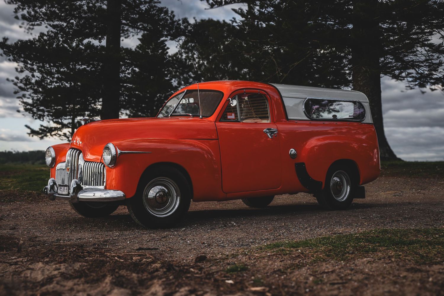 Vintage orange Holden FX ute parked on dirt road against backdrop of pine trees and moody sky.