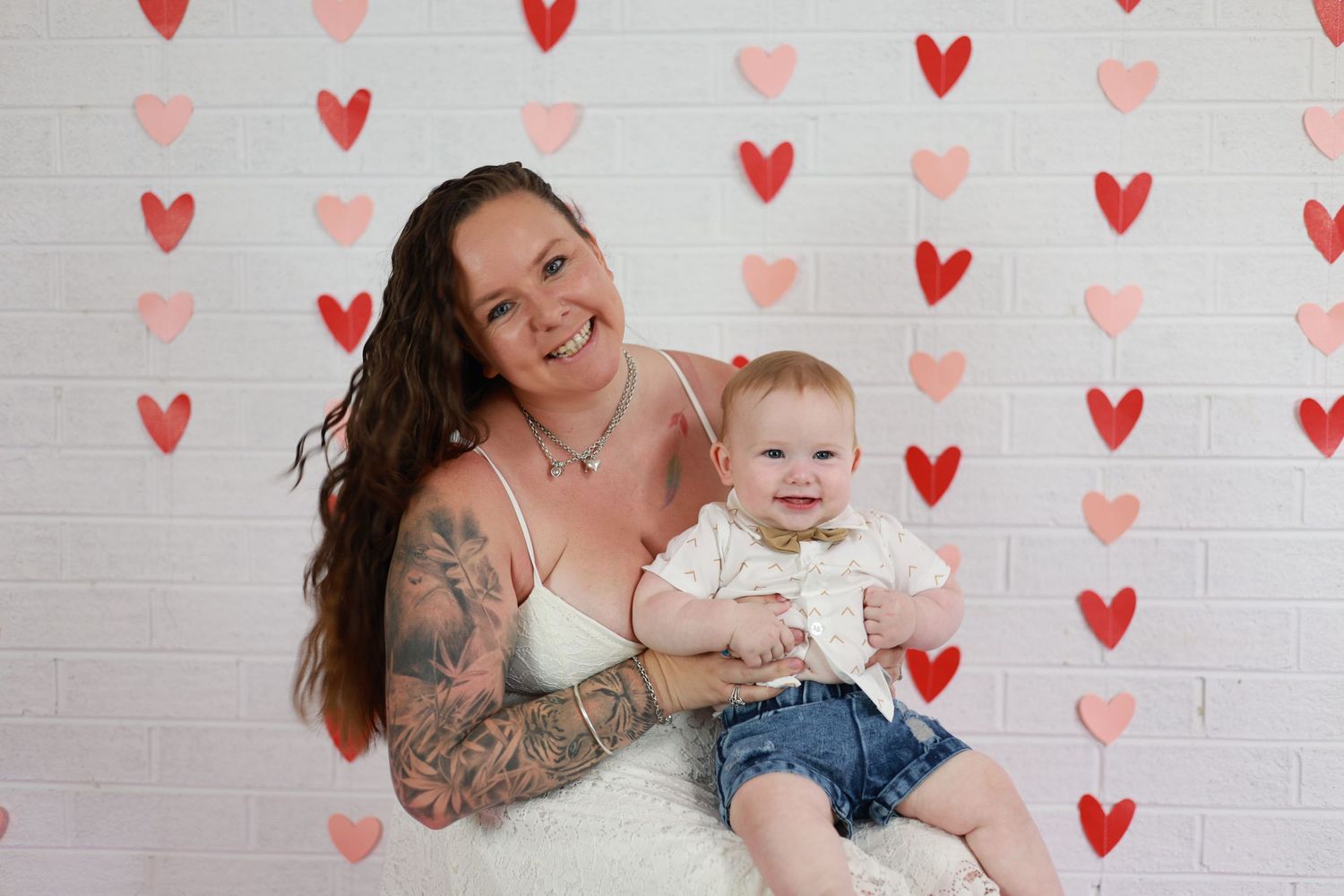 A parent and baby pose together against a white brick wall decorated with pink and red heart shapes.