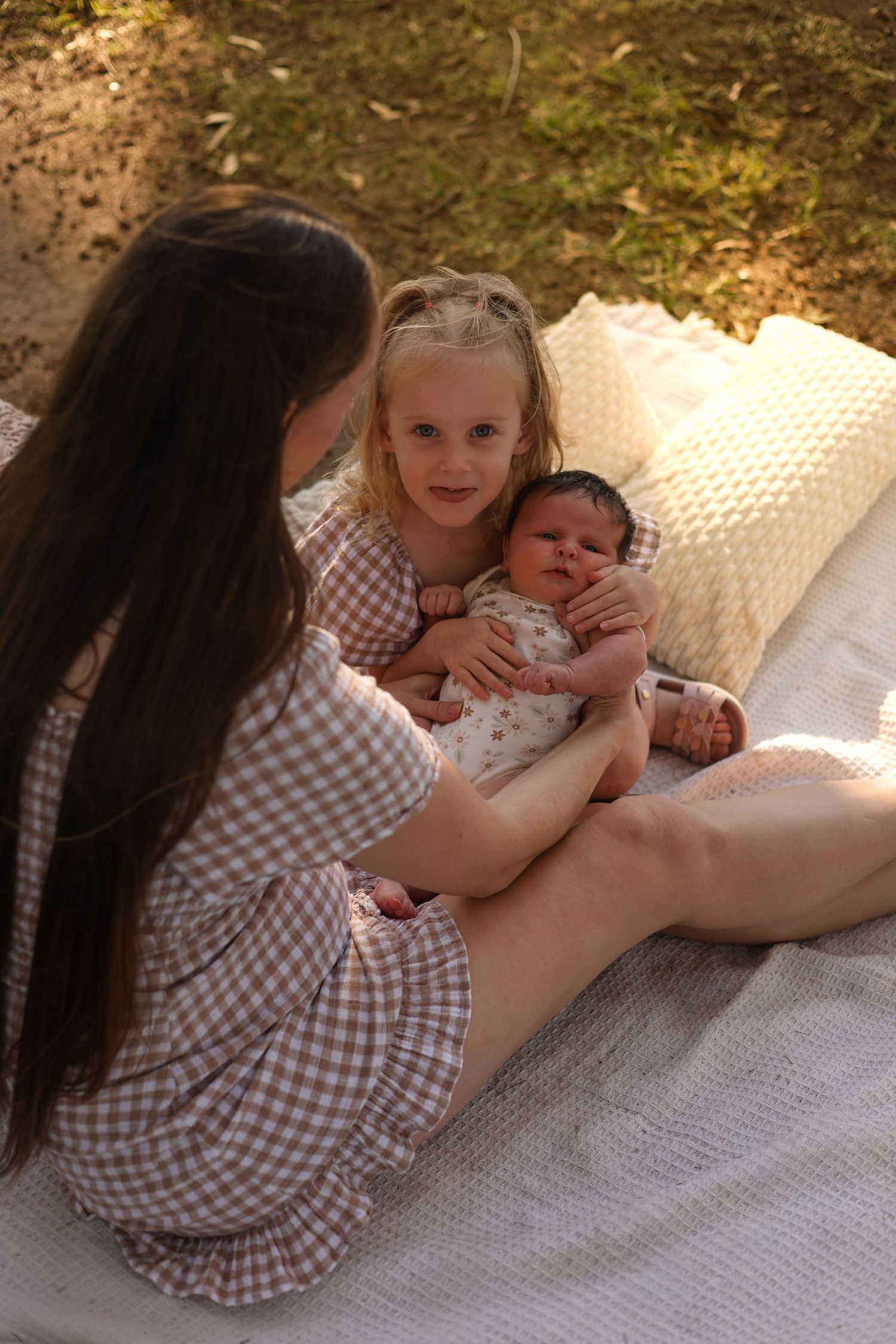 A mother sits on a white blanket outdoors holding a newborn baby with a young blonde girl smiling beside them.