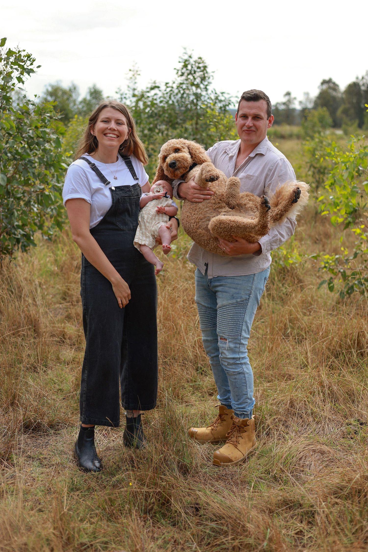 A young couple stands outdoors holding a baby and a golden doodle dog in a rural field setting.