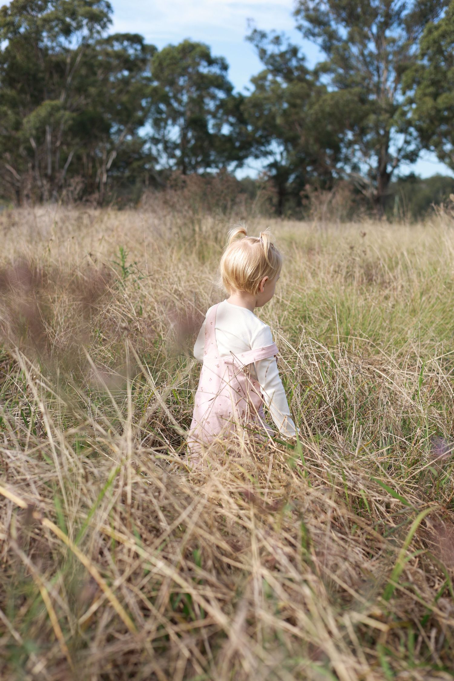 Young blonde girl in pink dress exploring tall grass field with trees in background on sunny day.