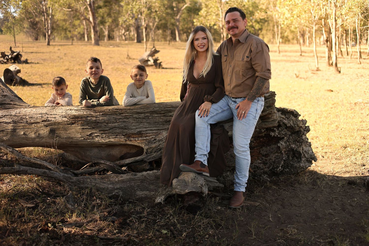 Family posing on a fallen log in a dry Australian bush setting during golden hour.