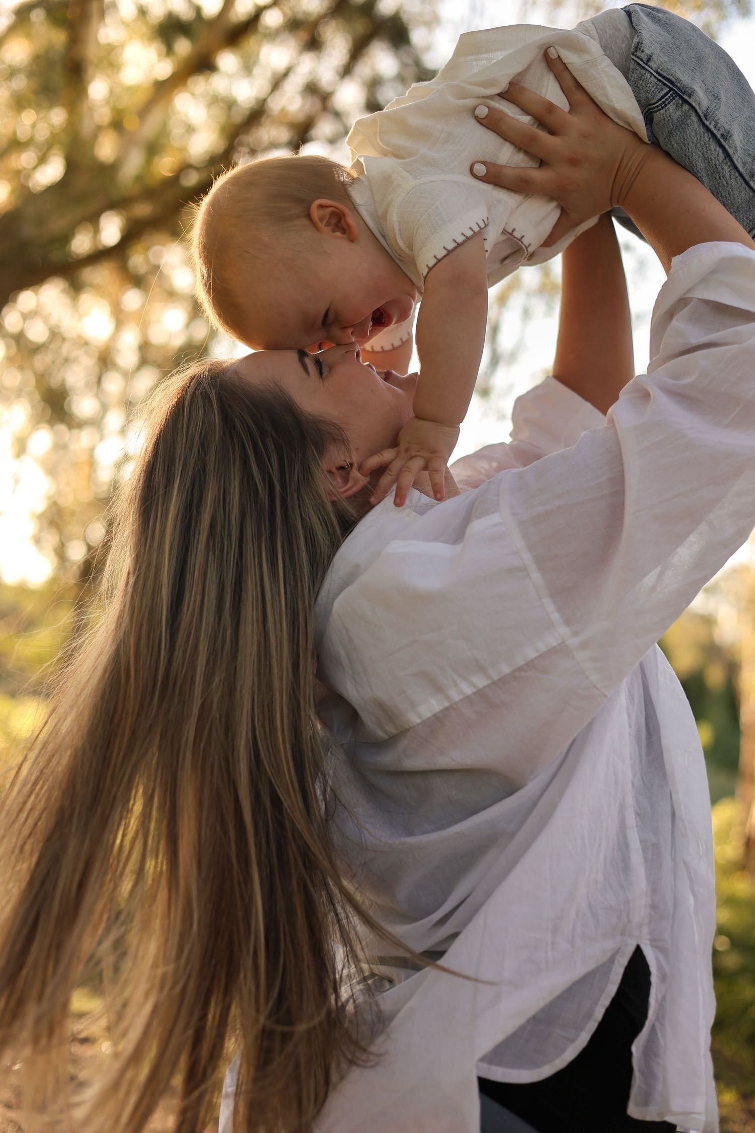 A mother joyfully lifts her baby in a sunlit park, both smiling in a tender outdoor moment.
