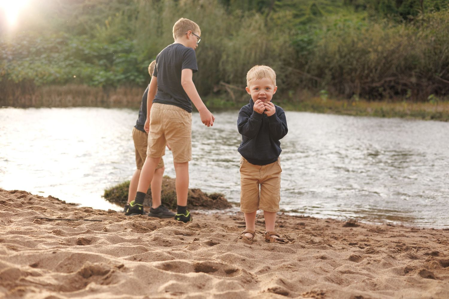 Two boys playing on a sandy riverbank surrounded by lush greenery on a sunny day.