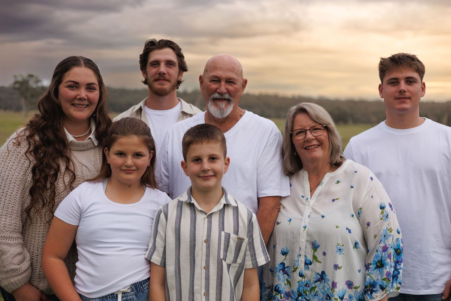 A multigenerational family of seven smiling together outdoors at sunset in a rural field setting.
