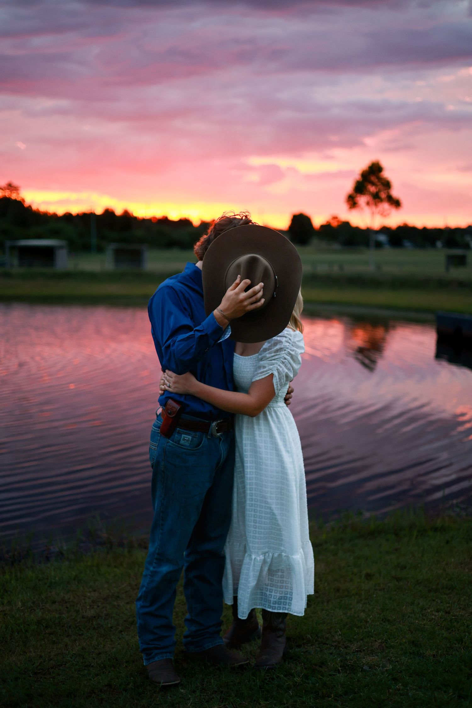 A couple embraces by a pond during a vibrant pink and orange sunset, creating a romantic silhouette against the colorful sky.