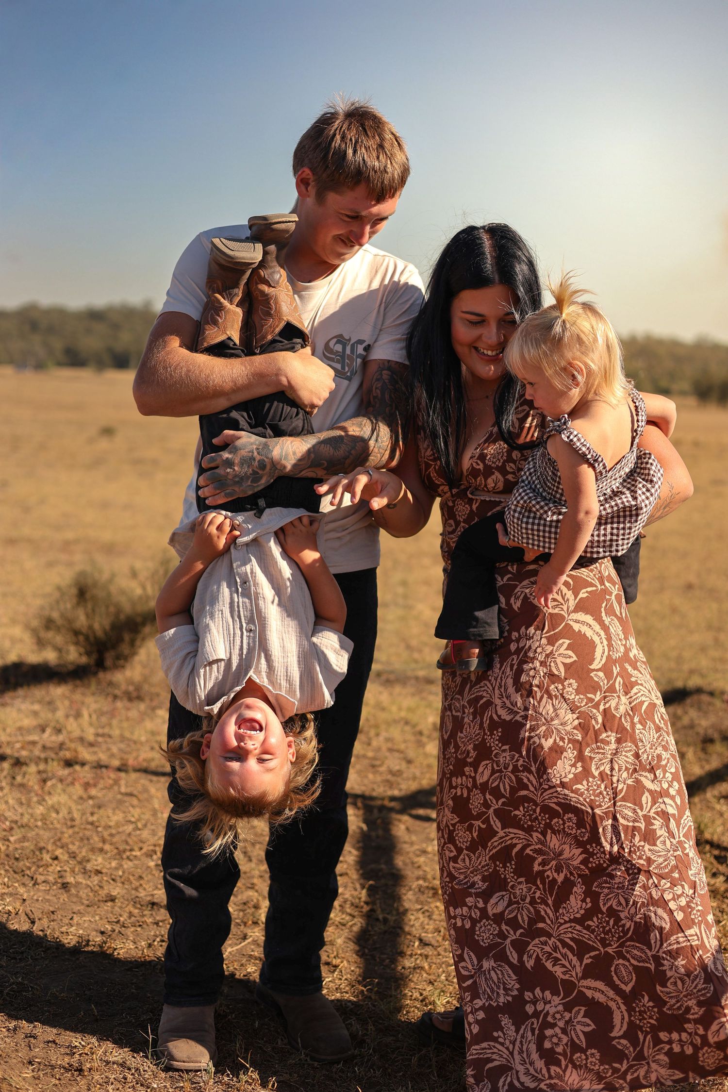 Happy family with tattooed dad holding toddler upside down while mom carries baby in sunny field.