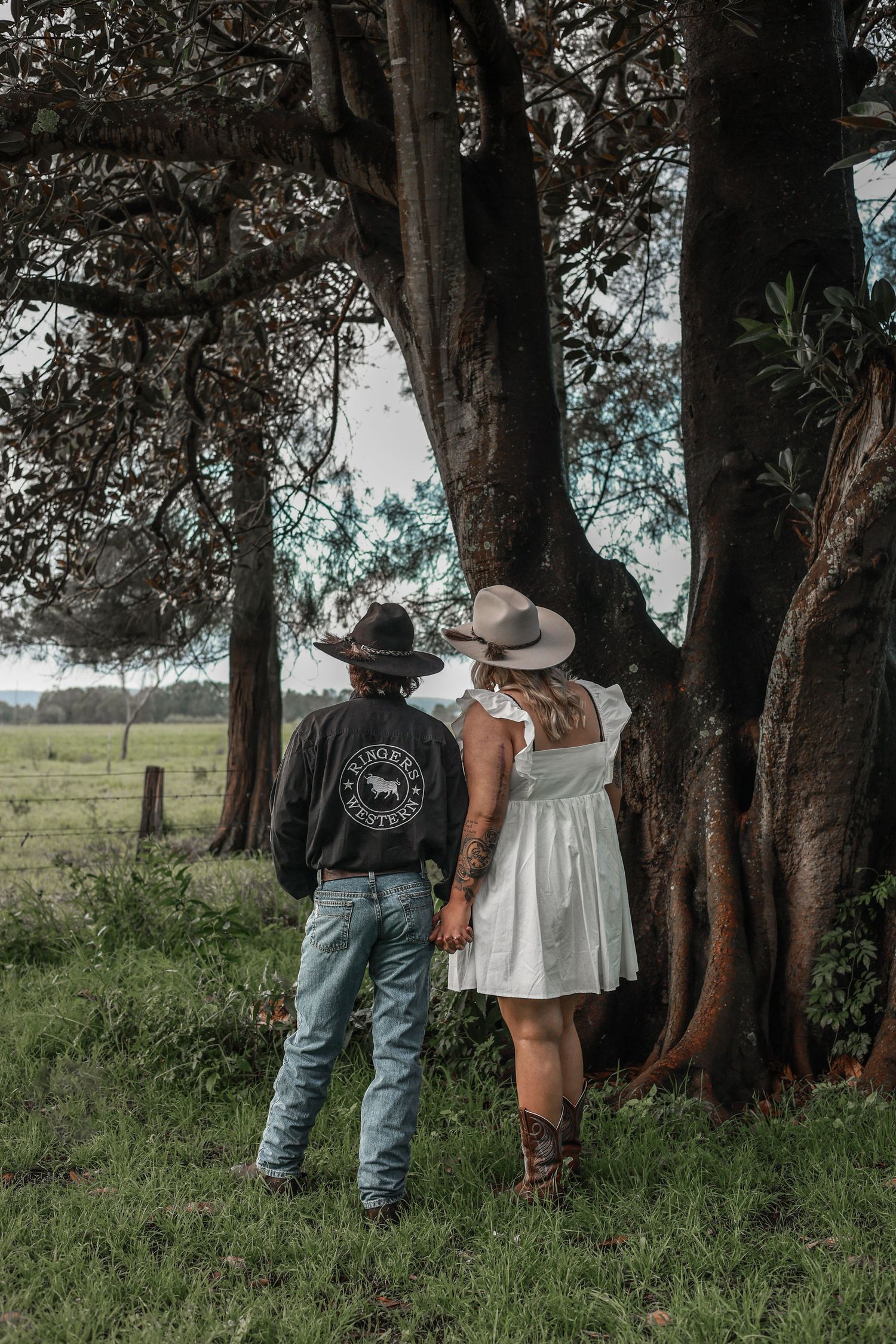 A couple standing together under large shady trees in a rustic outdoor setting.