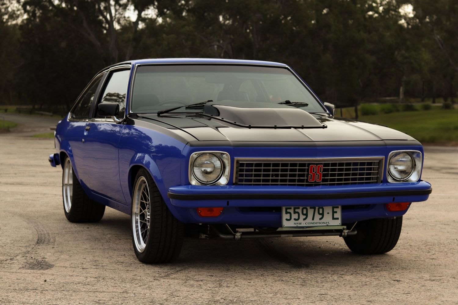 A classic blue Holden Torana with chrome wheels parked on gravel with trees in the background.