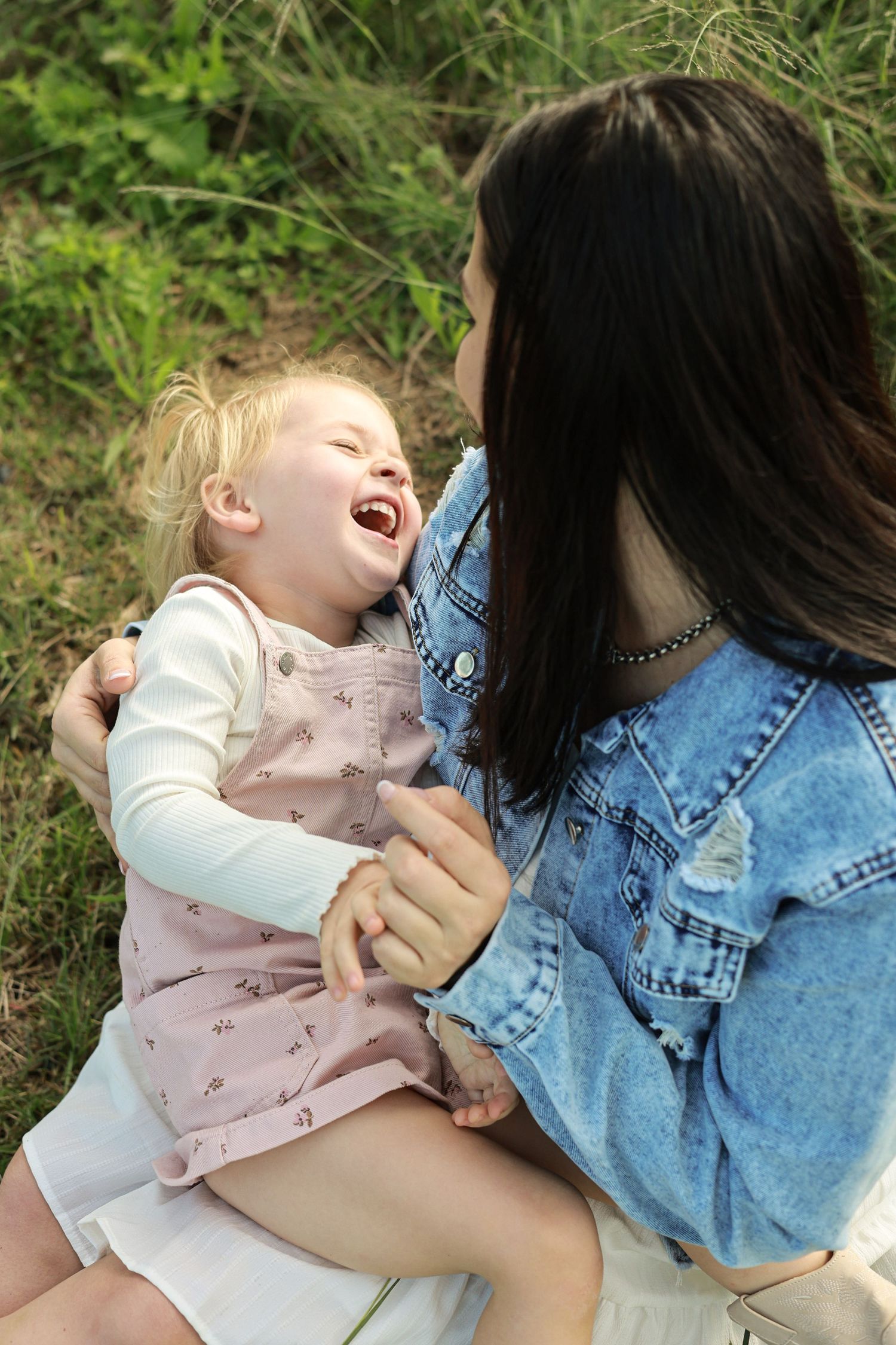 A joyful toddler laughing while being held by a dark-haired woman in a denim jacket outdoors.