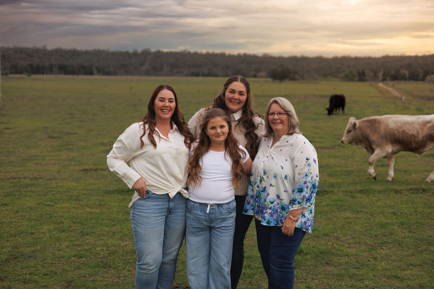 Four women smile together in a green pasture at sunset with cattle grazing in the background.