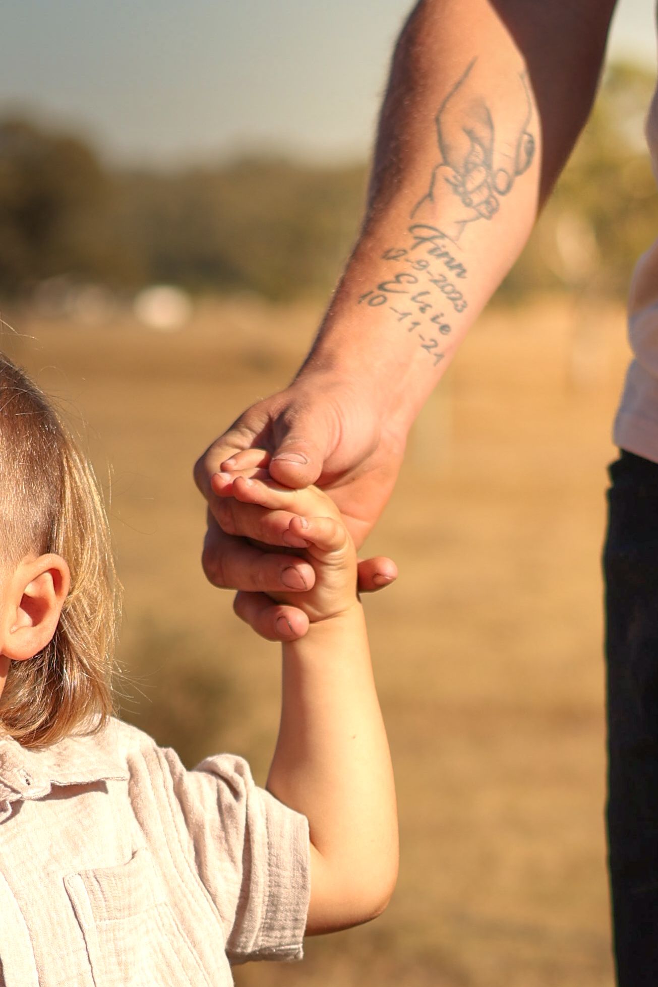 Tattooed adult hand holding a small child's hand outdoors in a sunny field.