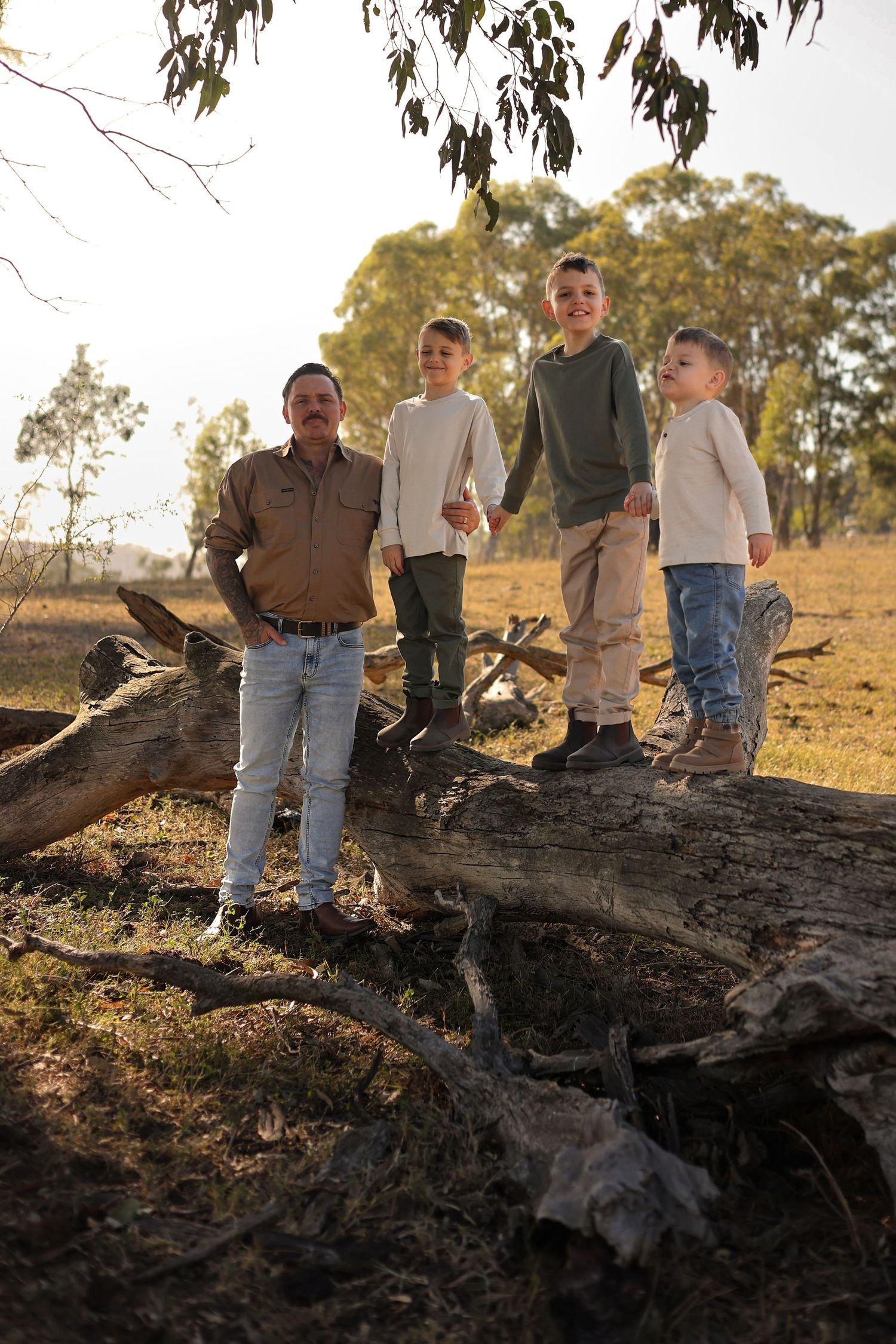 A man and three boys stand on a fallen log in a sunlit rural Australian landscape with eucalyptus trees.