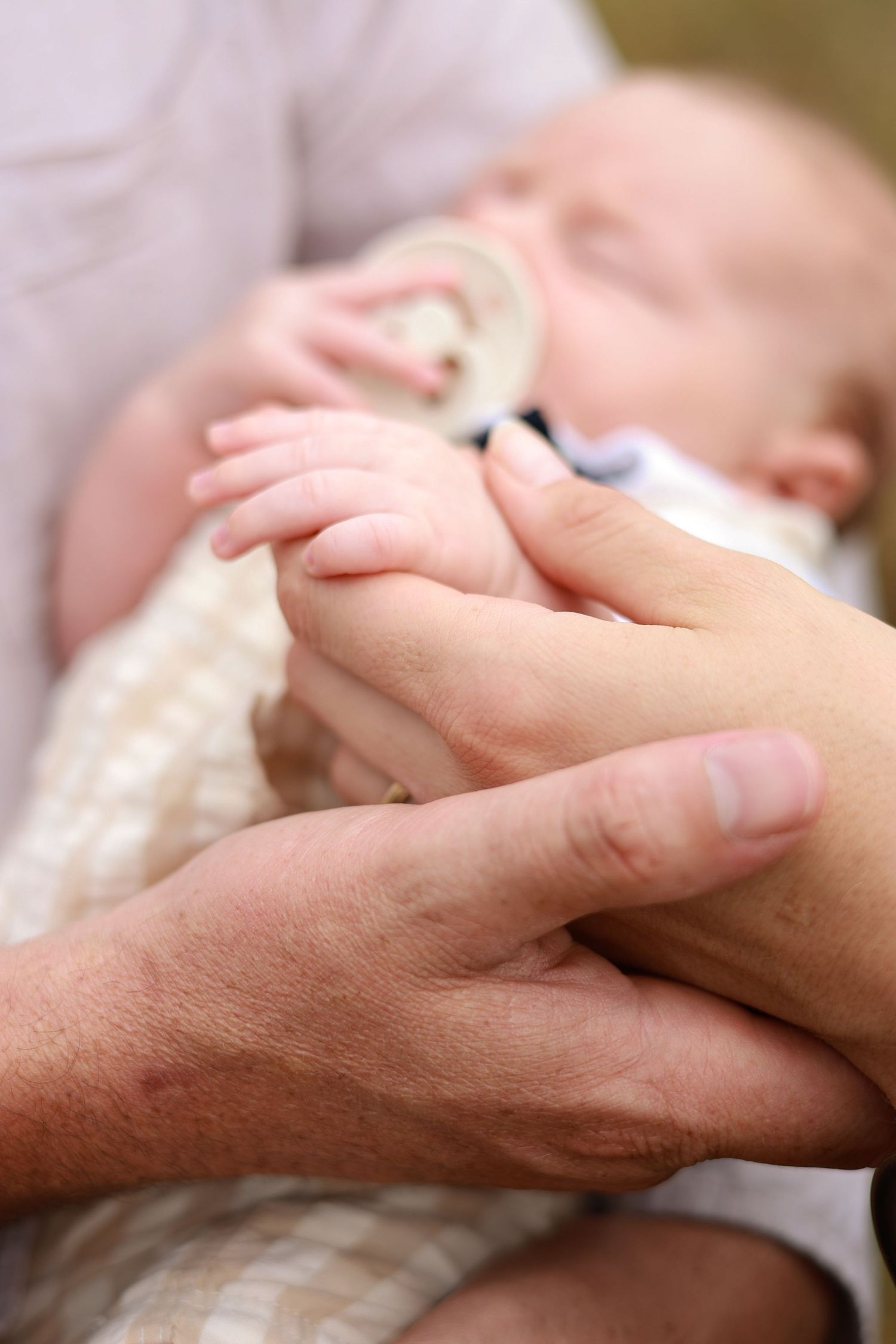 A newborn baby with a pacifier gently holds an adult's finger while being cradled in loving arms.