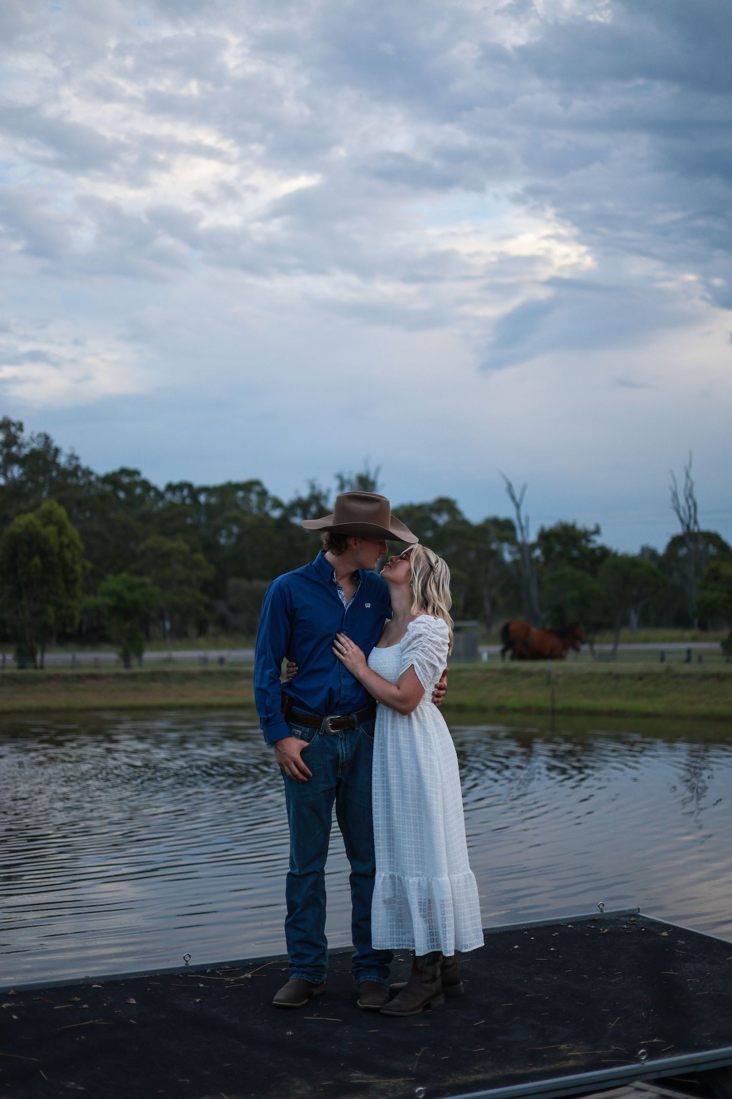 A couple stands together on a dock by a lake at dusk with moody clouds overhead.