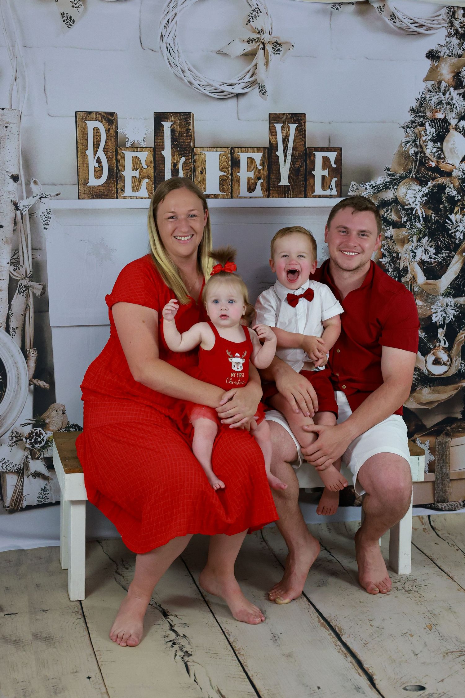 Family Christmas portrait in matching red outfits seated on bench with 'BELIEVE' sign and decorated tree in background.