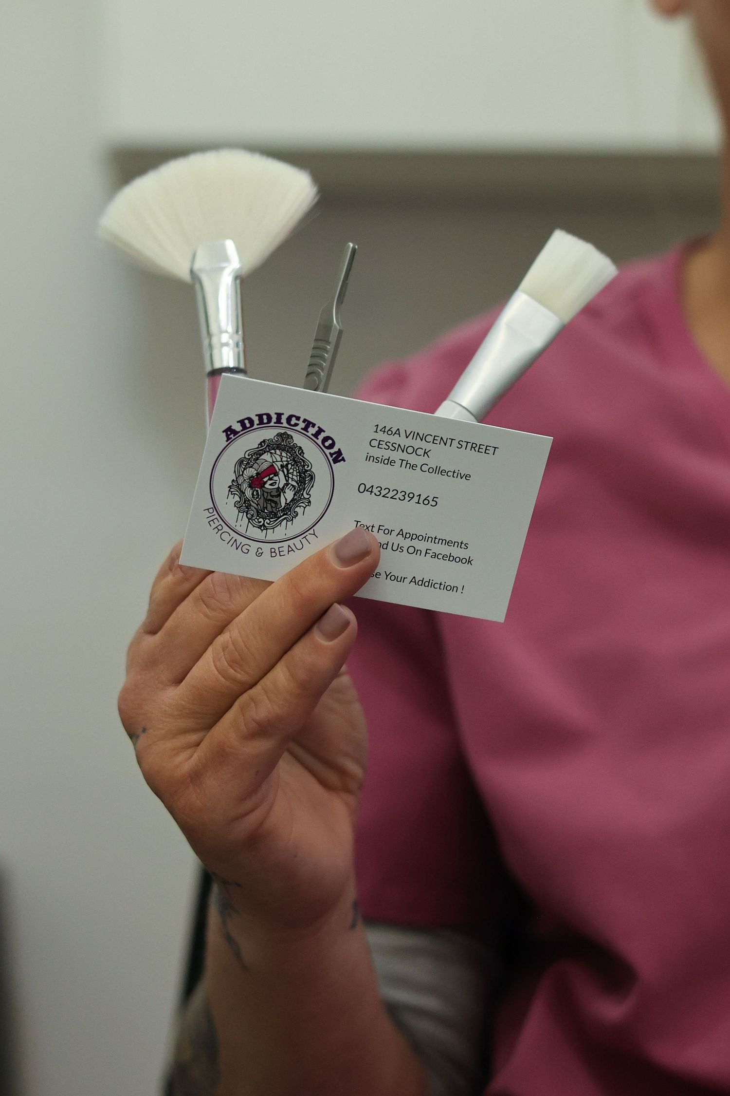 Person in pink scrubs holding makeup brushes and an Addiction Tattooing & Beauty business card showing address and contact details.