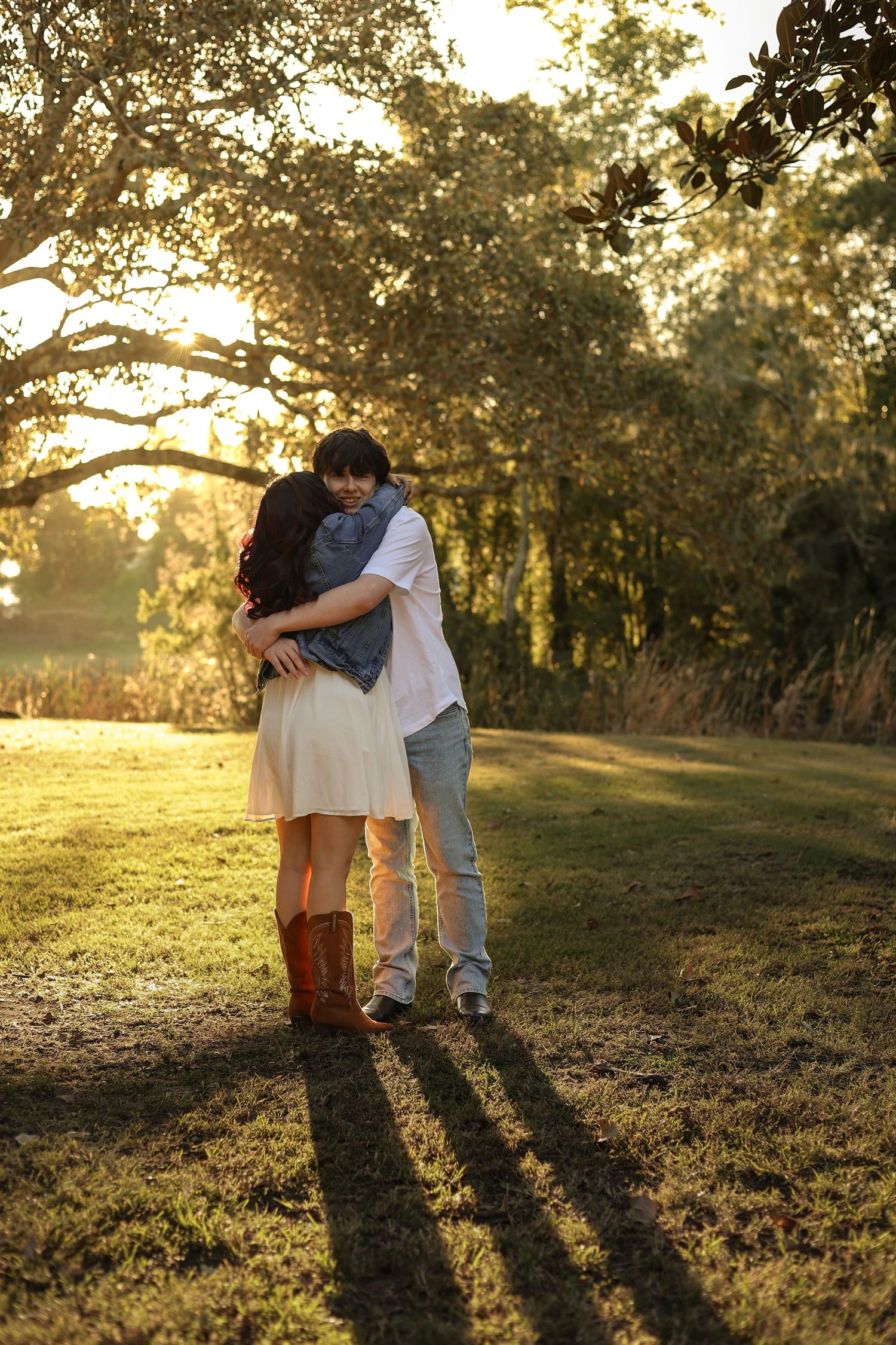 Couple embracing at golden hour in a sunlit park, woman in white dress and red boots, man in casual attire.