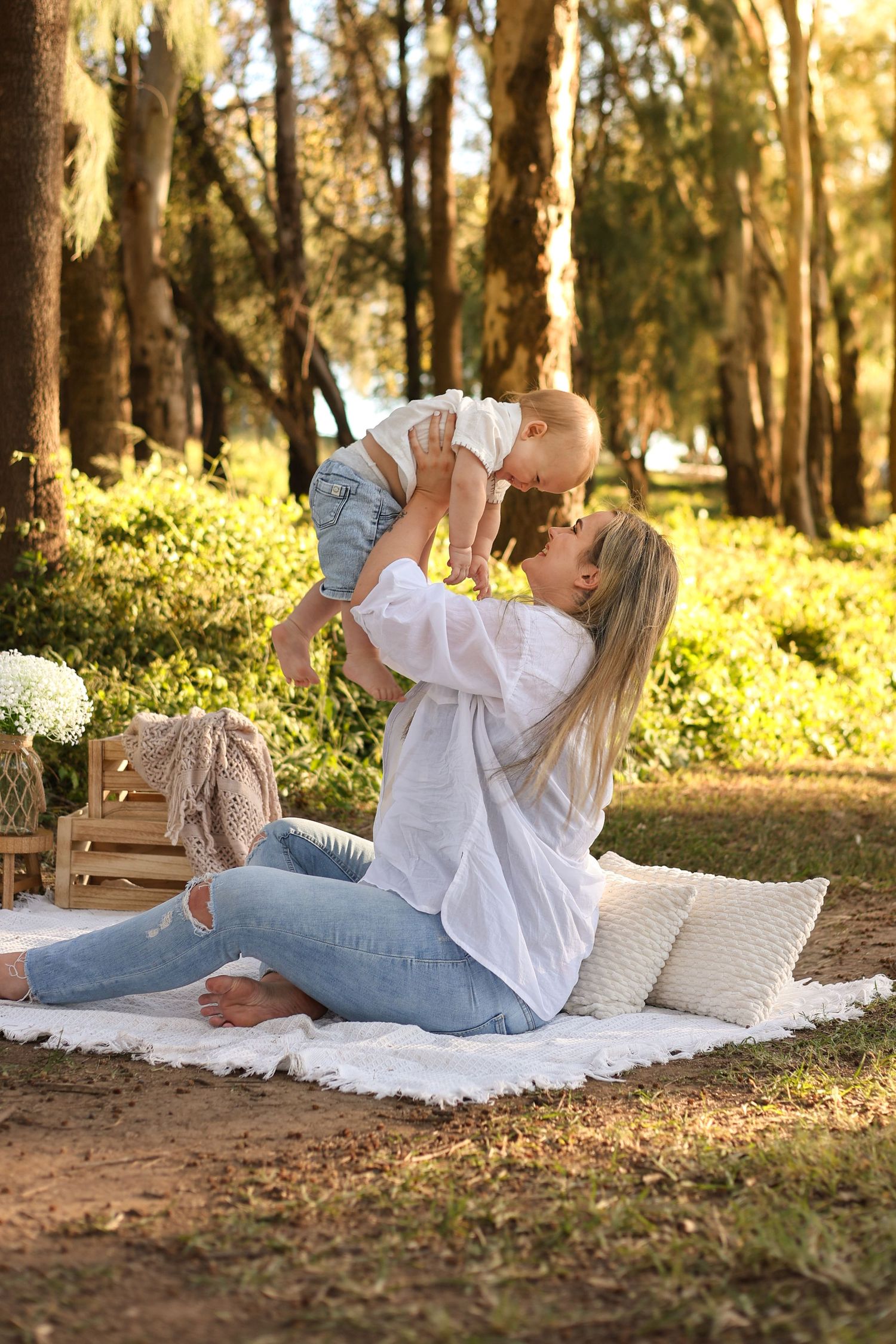 A mother sits on a white blanket in a sunny park, lovingly lifting her baby up while smiling outdoors.