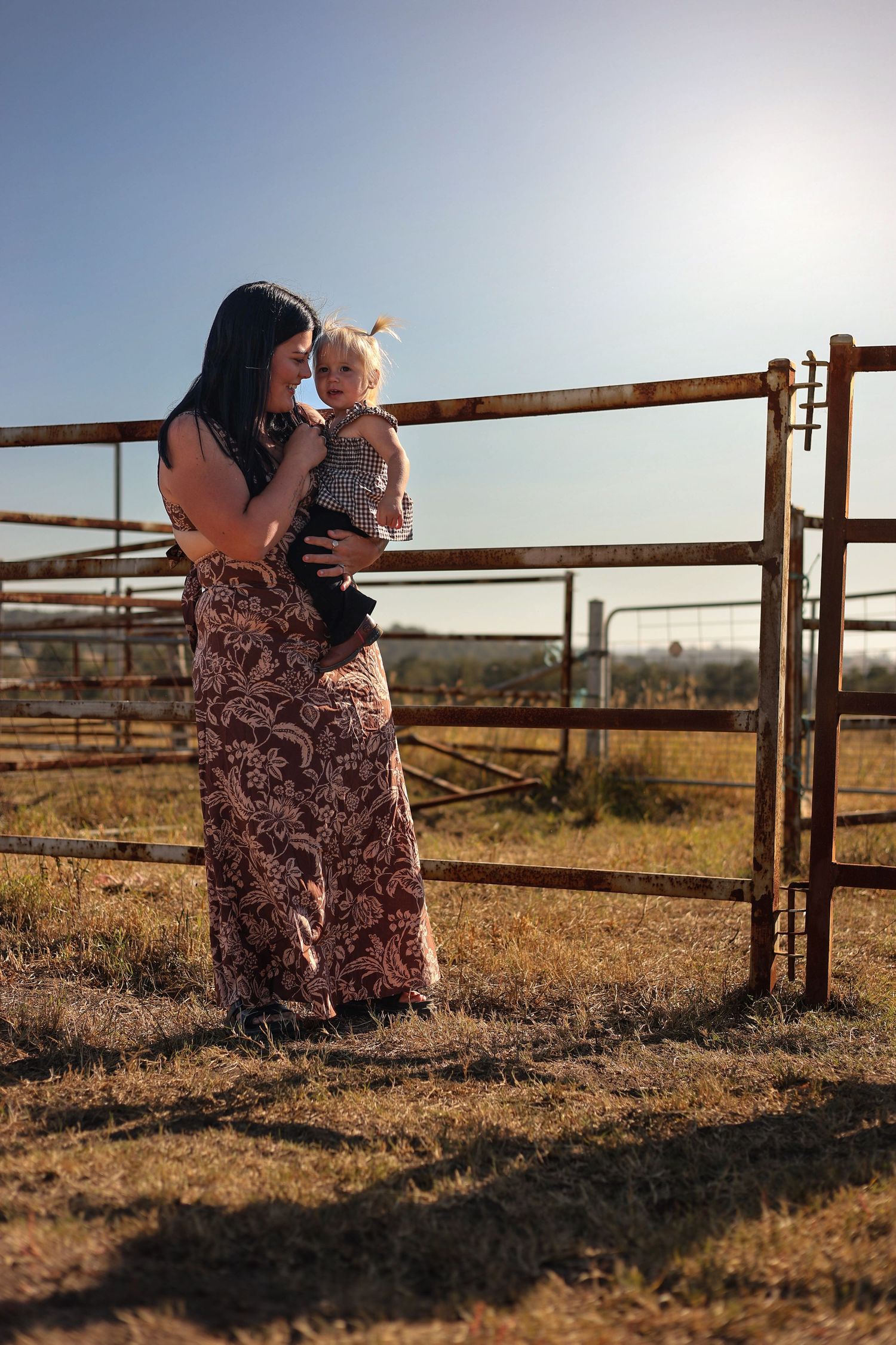A woman in a floral maxi dress holds a toddler near a rustic farm gate in golden sunlight.