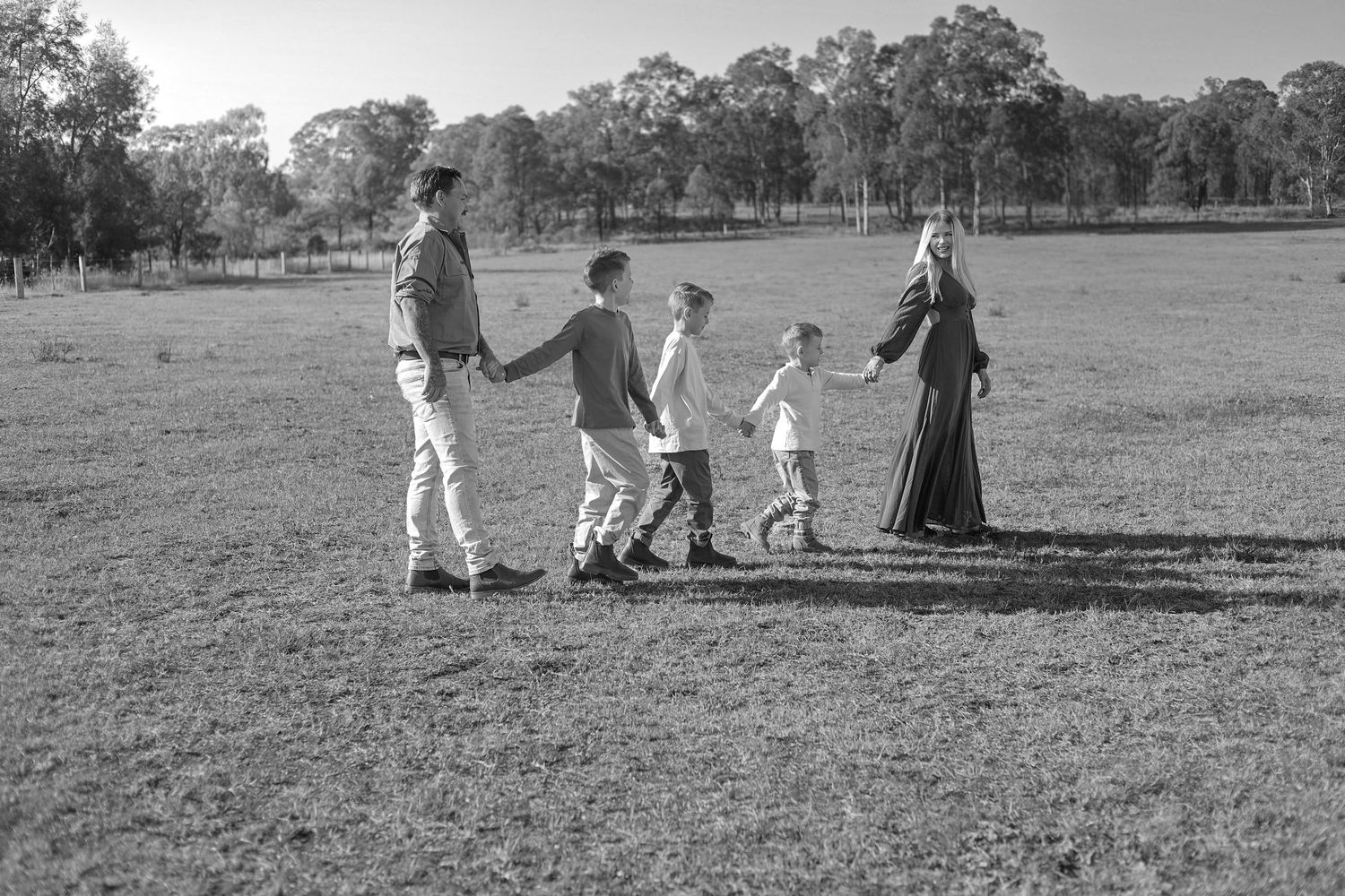 Black and white photo of a family holding hands walking through a rural field with trees in the background.