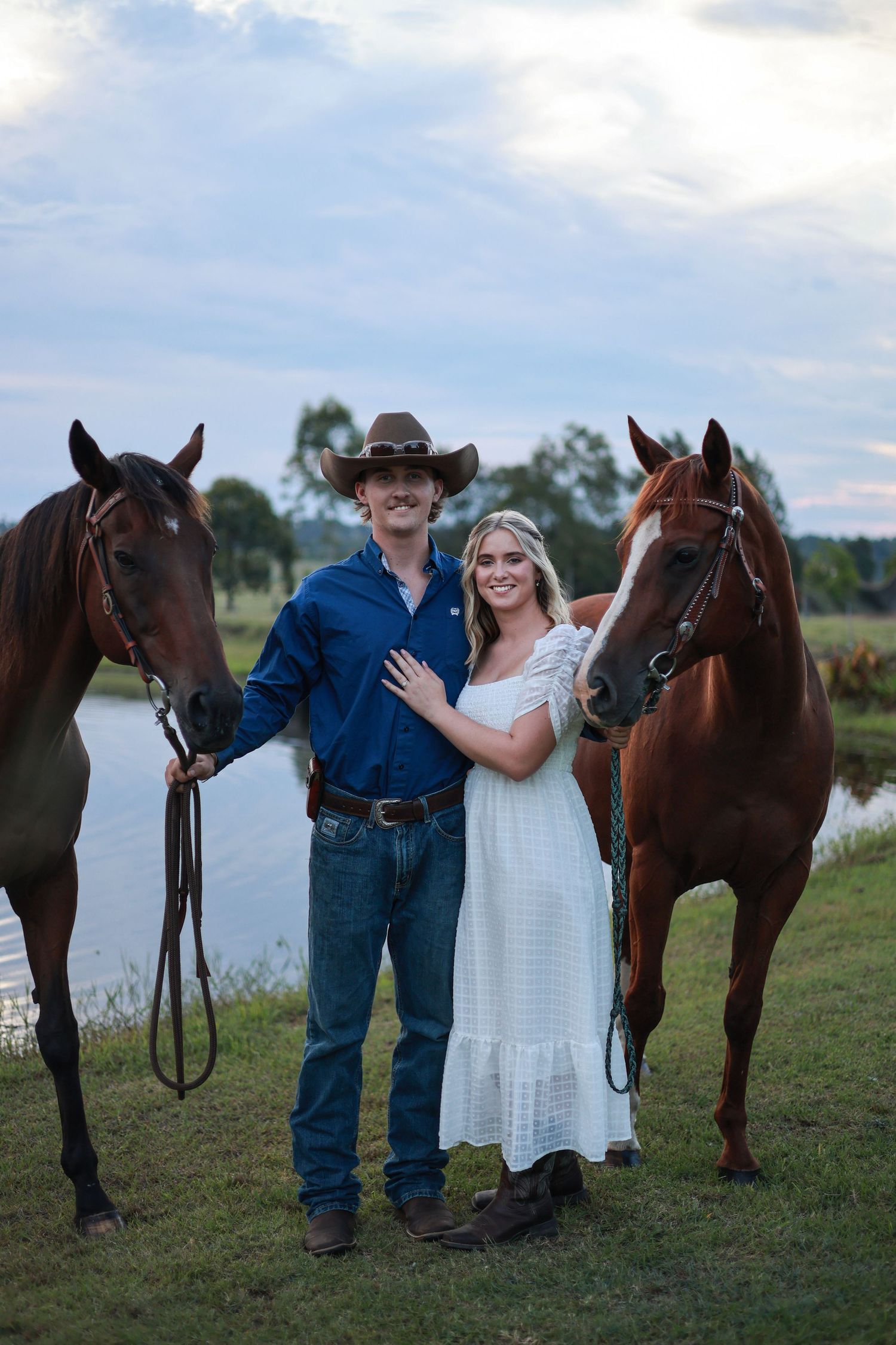 A couple in western attire stands with their horses by a lake during a rustic outdoor photoshoot.
