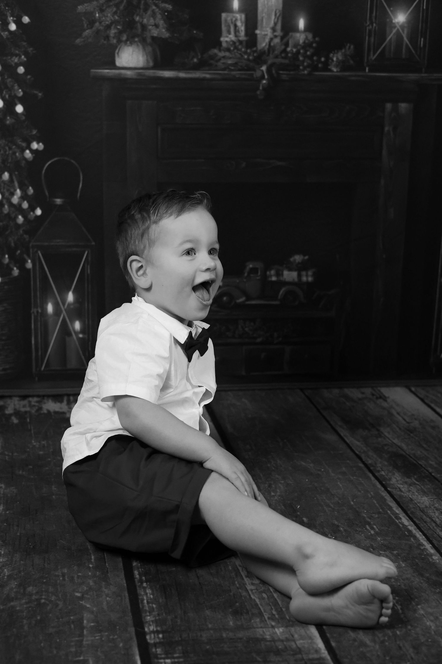 Black and white photo of an excited child in formal attire sitting on a wooden floor with lantern decorations in background.