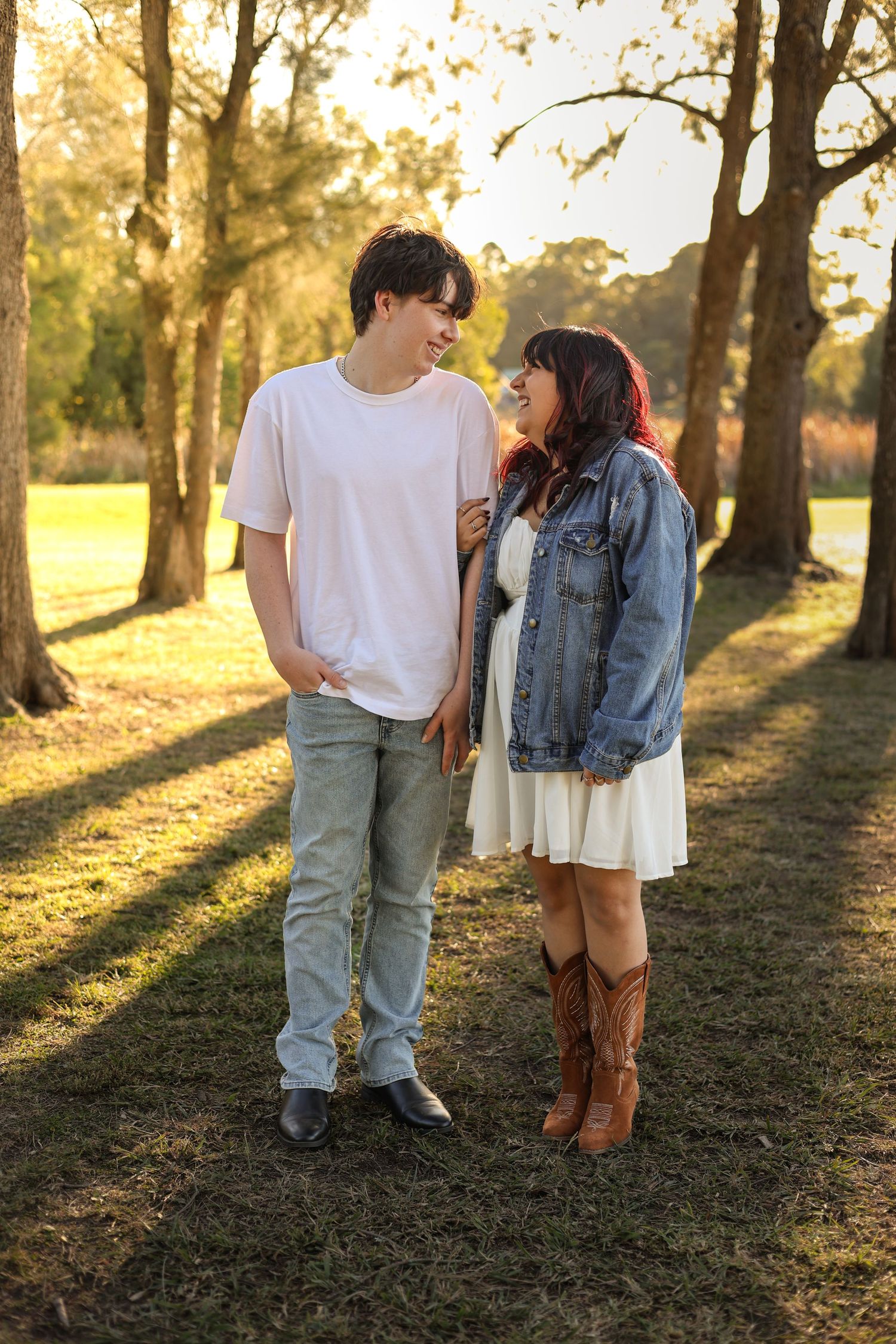 A young couple smiles at each other in a sunlit park, she wears a denim jacket and cowboy boots.