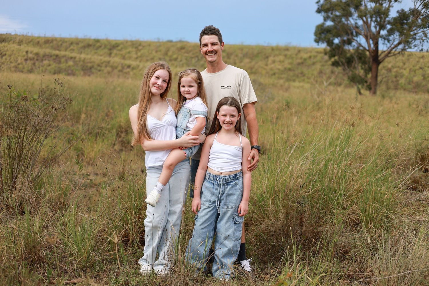 A smiling family of four poses in a grassy field, with a man holding a young girl flanked by two older girls.