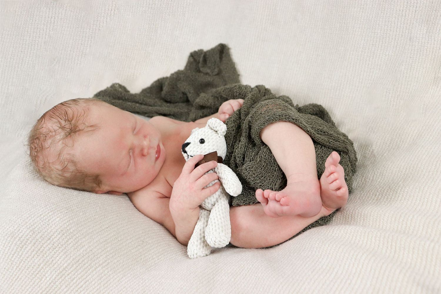 A sleeping newborn wrapped in an olive green blanket cuddles with a small white stuffed toy bear on a beige backdrop.