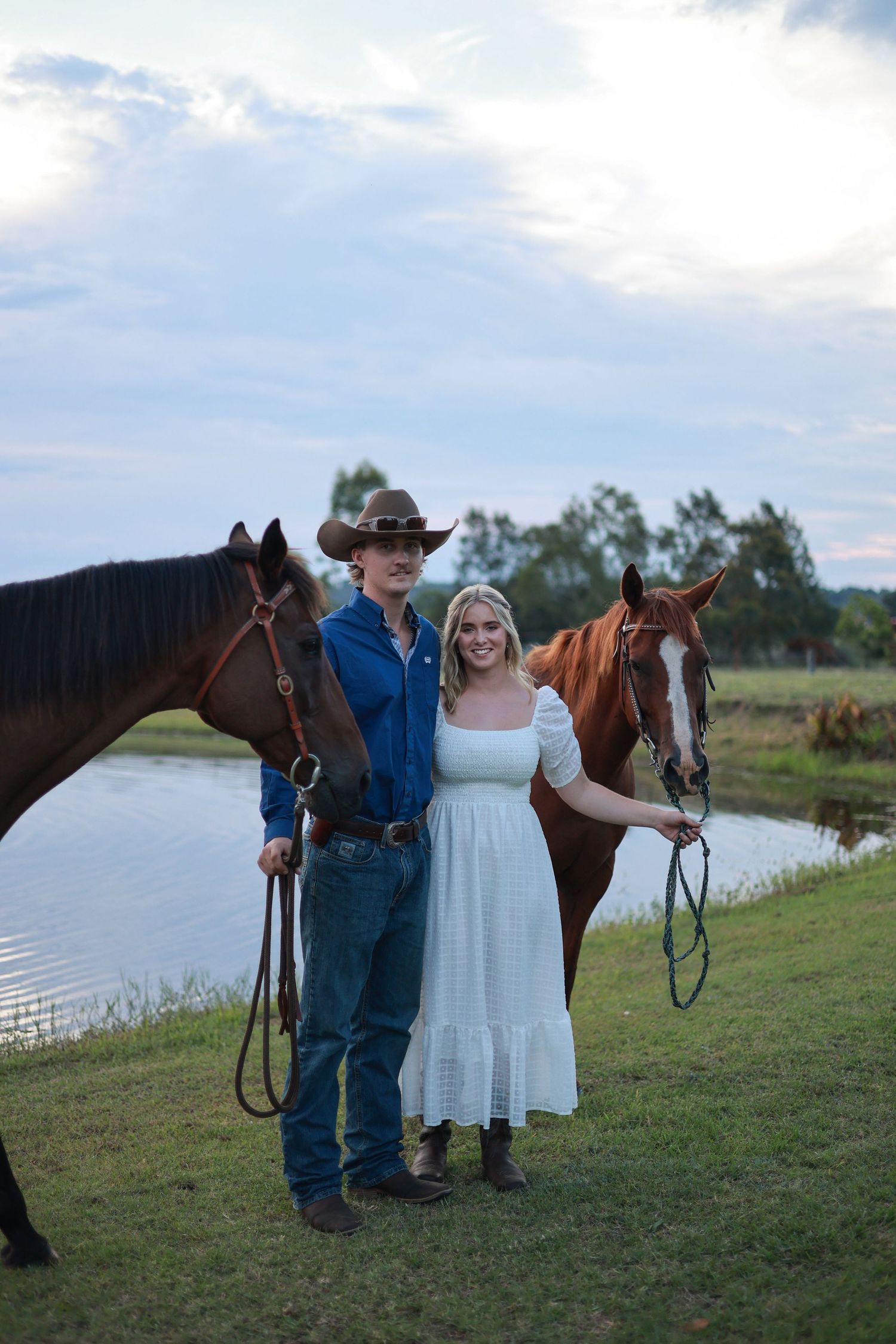A couple stands with two horses by a lake, the sun setting behind them in a rural countryside setting.