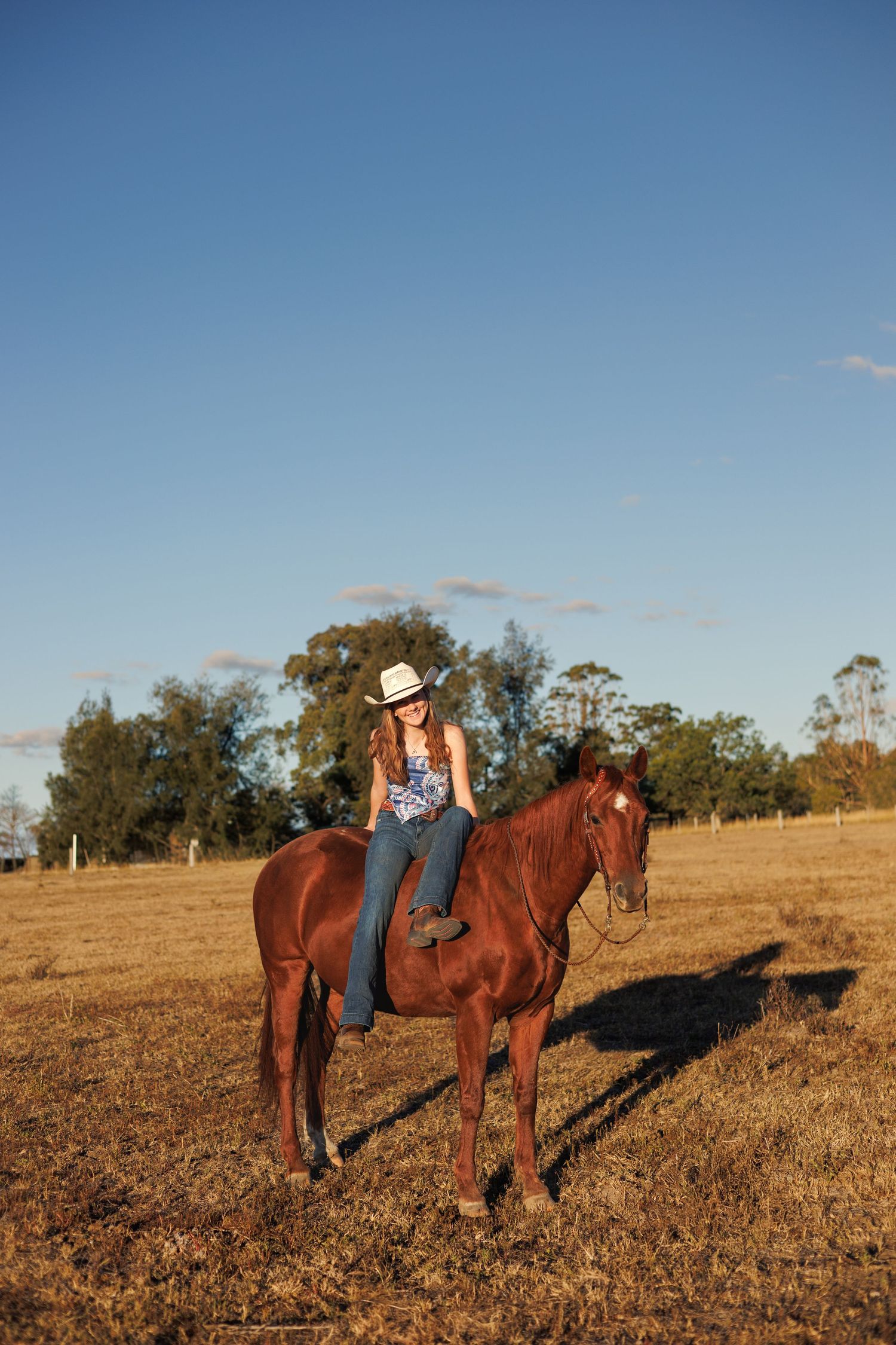 Young woman in cowboy hat riding a chestnut horse in a sunny rural field with blue sky backdrop.