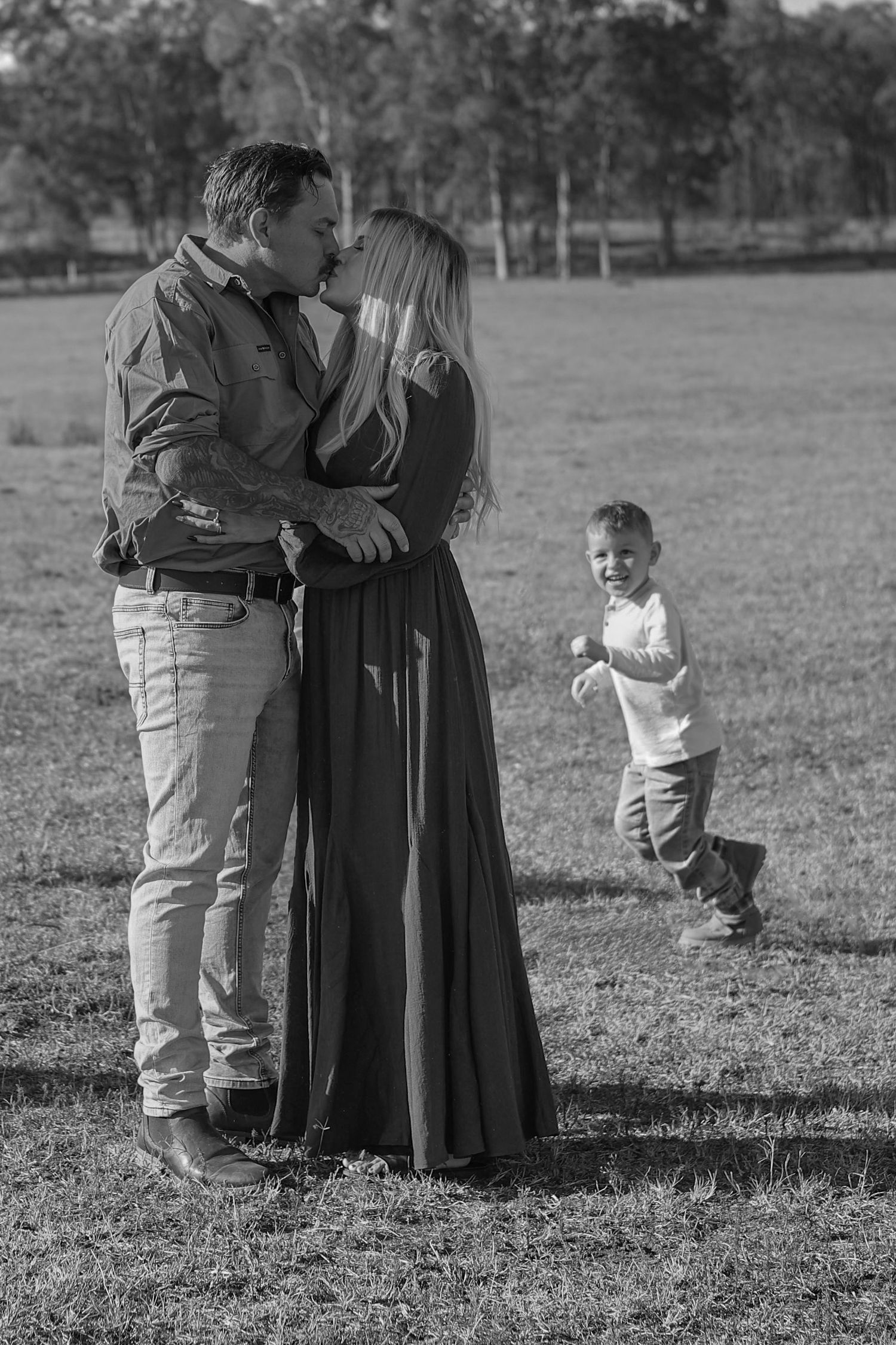 Black and white photo of a couple kissing in a field while a young child plays behind them.