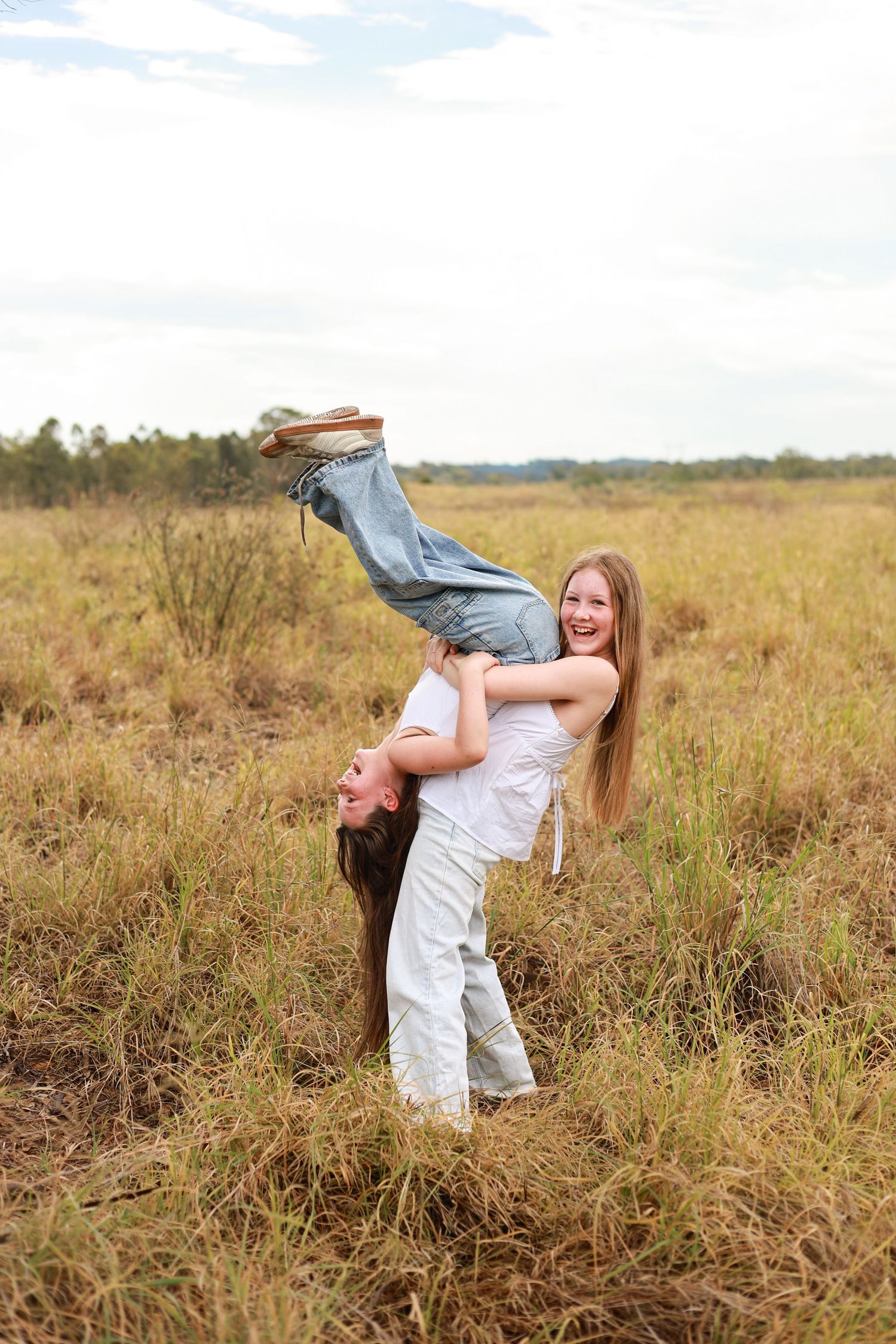 Two young women laughing and playing in a field, one lifting the other upside down outdoors.