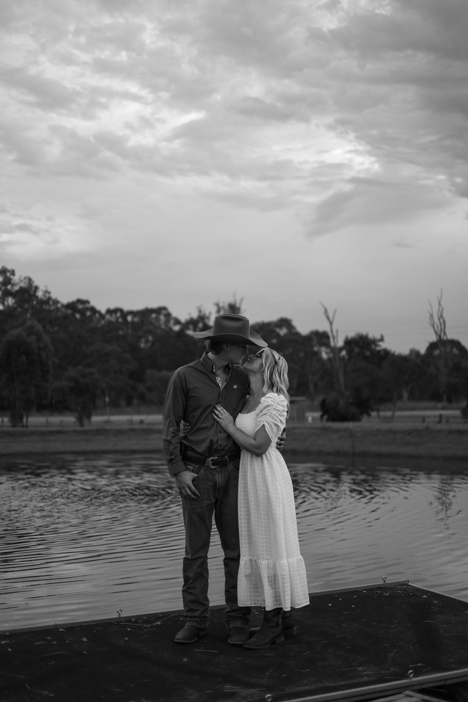A romantic black and white photo series of a couple standing on a wooden dock by a serene lake at dusk.