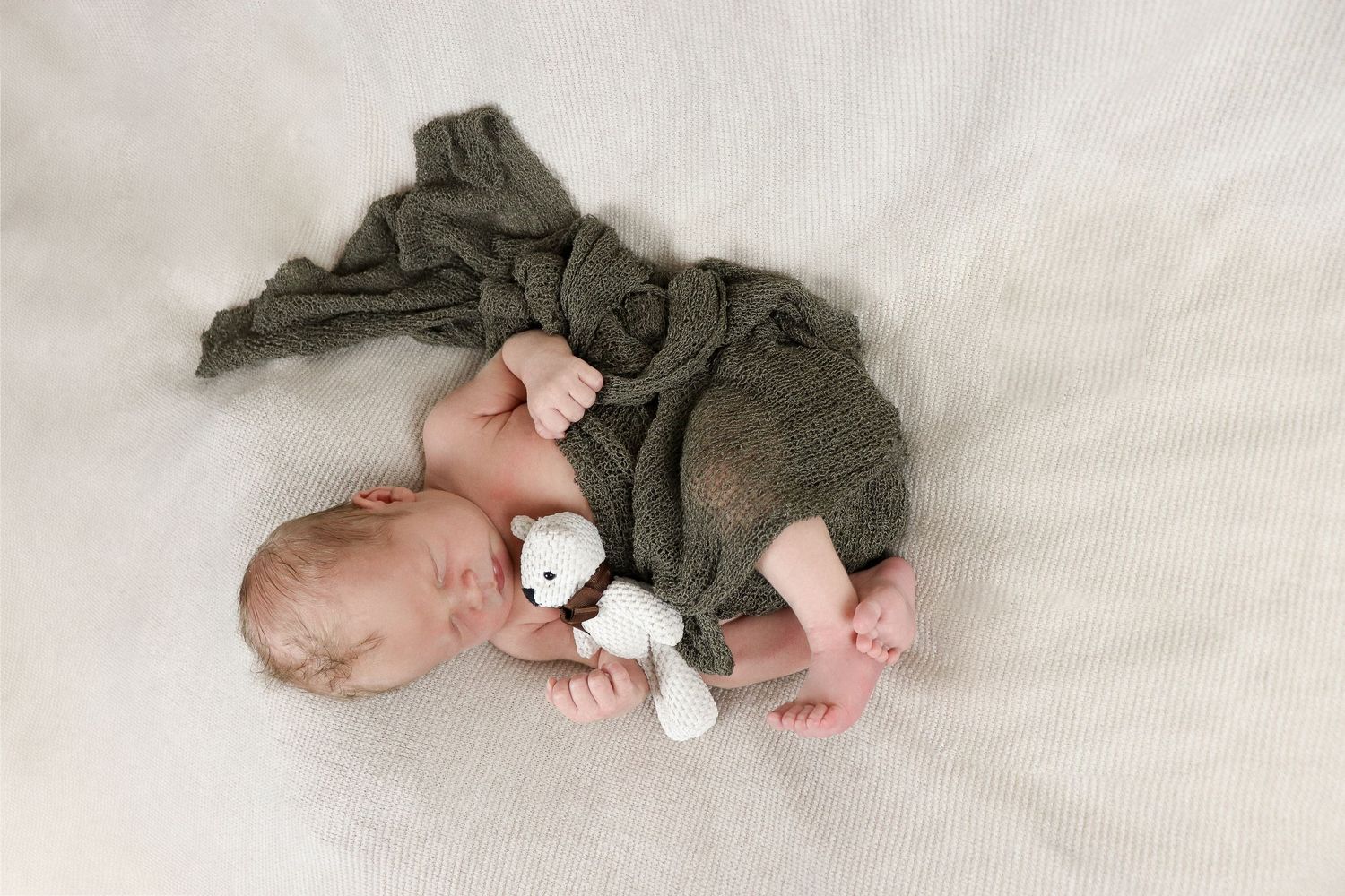 Small baby sleeping wrapped in cozy green knit swaddle with plush bear toy companion on soft white surface.