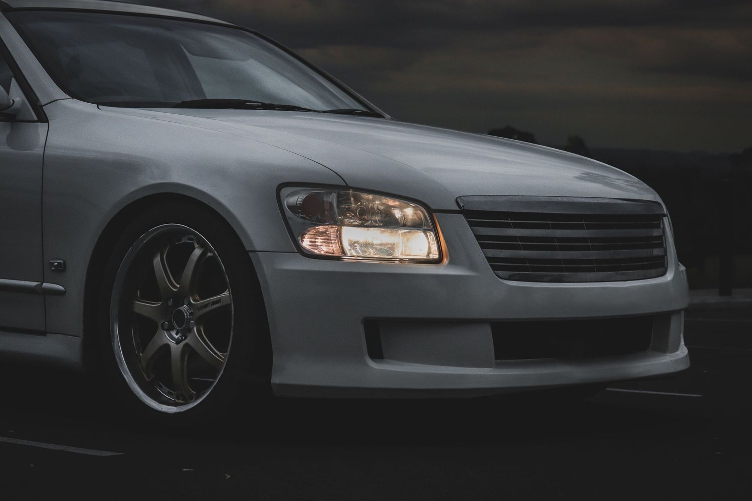 Front corner view of a white M35 stagea wagon with chrome & bronze wheels and headlights in moody lighting.