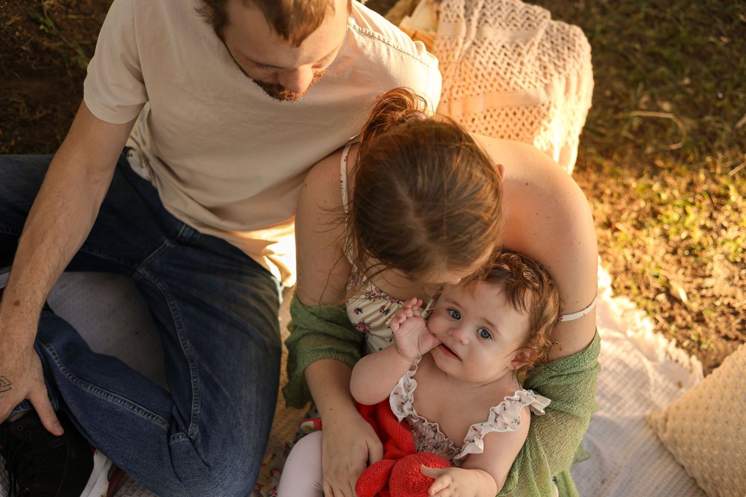 A young family sitting outdoors on a blanket, parents lovingly holding their baby girl in warm sunlight.