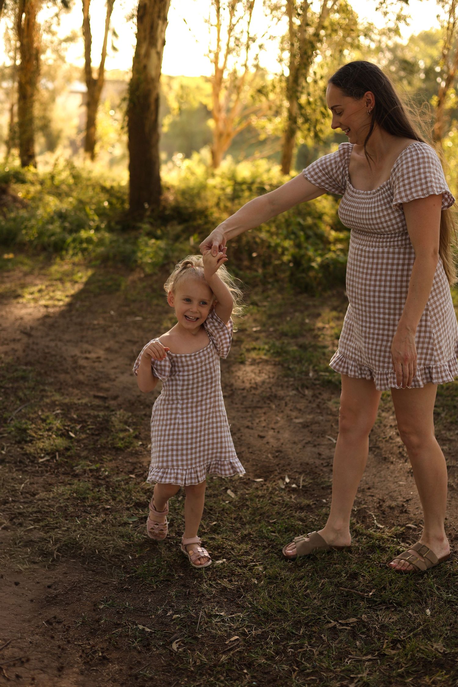 Mother and daughter in matching gingham dresses hold hands on a sunlit forest path at golden hour.