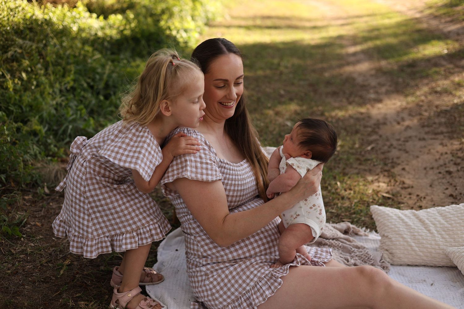 A mother in a gingham dress sits outdoors holding a newborn while her young blonde daughter in matching dress looks on.