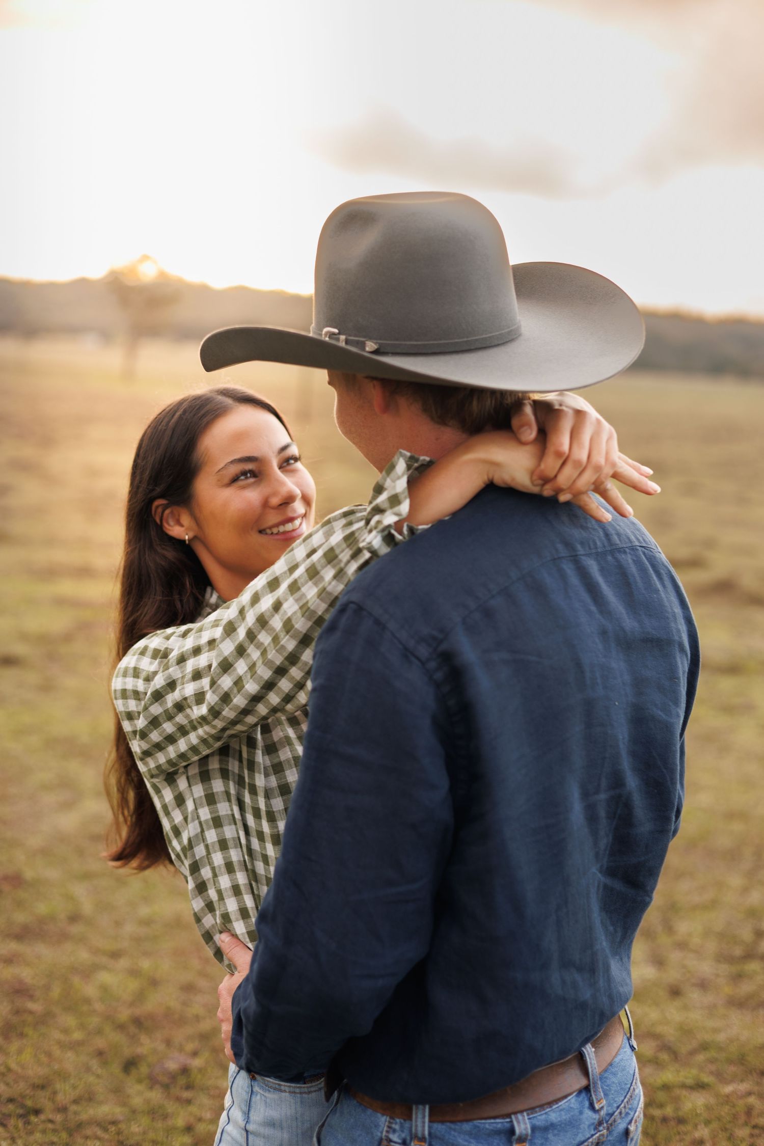 A couple embraces in a golden field at sunset, woman smiling as man in cowboy hat holds her close.