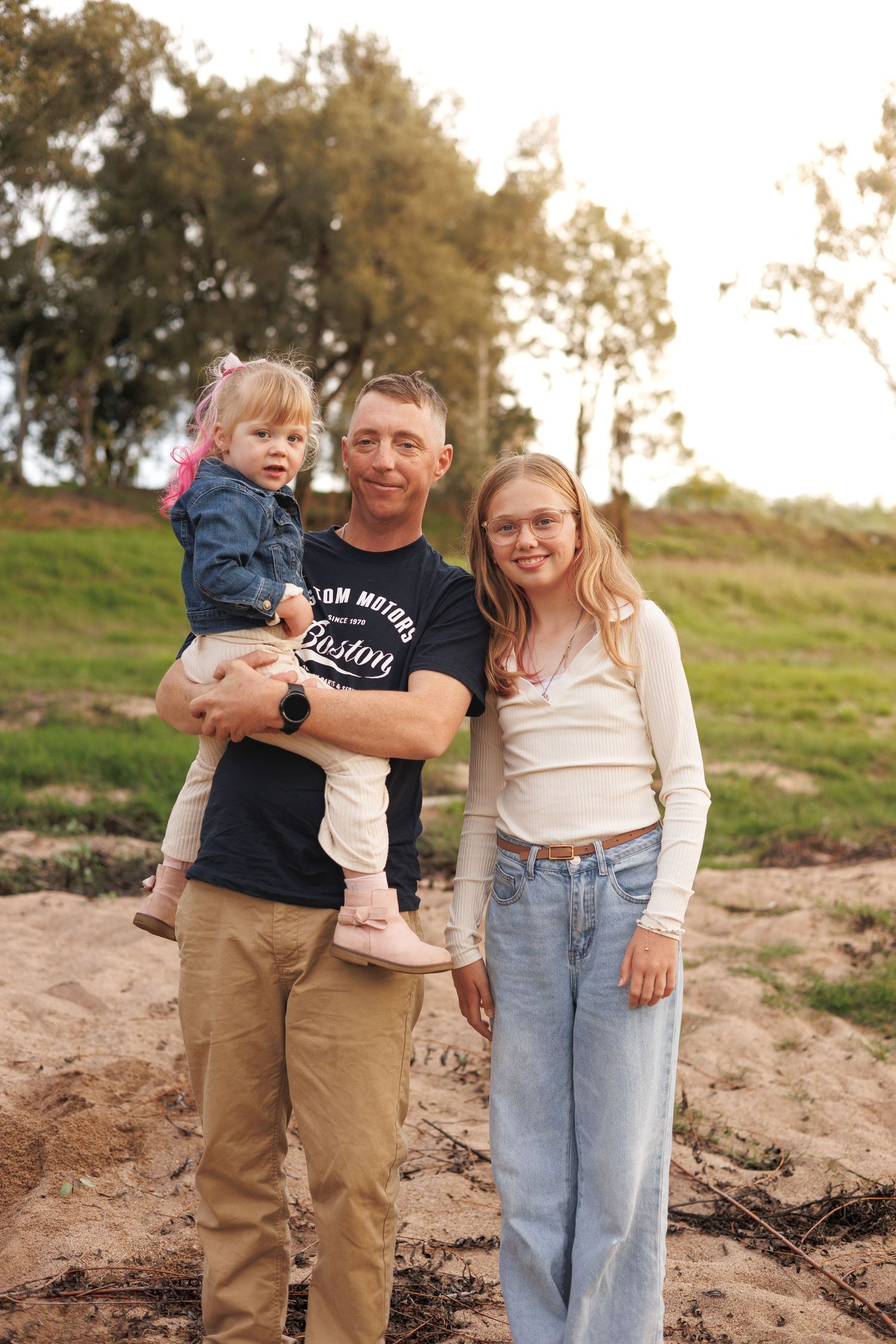 A man and young woman smile outdoors holding a toddler girl on sandy ground with trees behind them.