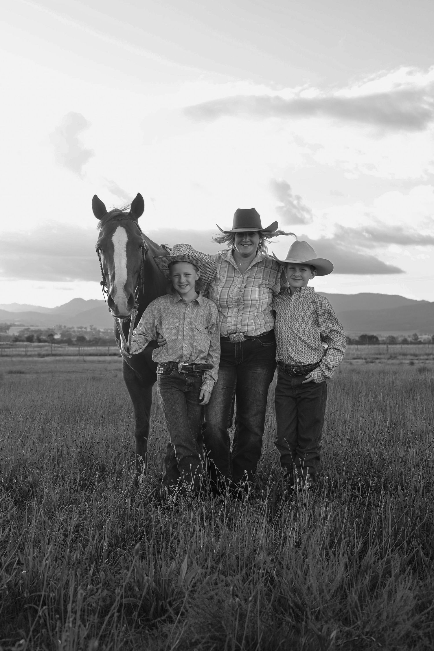 Black and white photo of a woman and two children in cowboy hats posing with a horse in an open field.