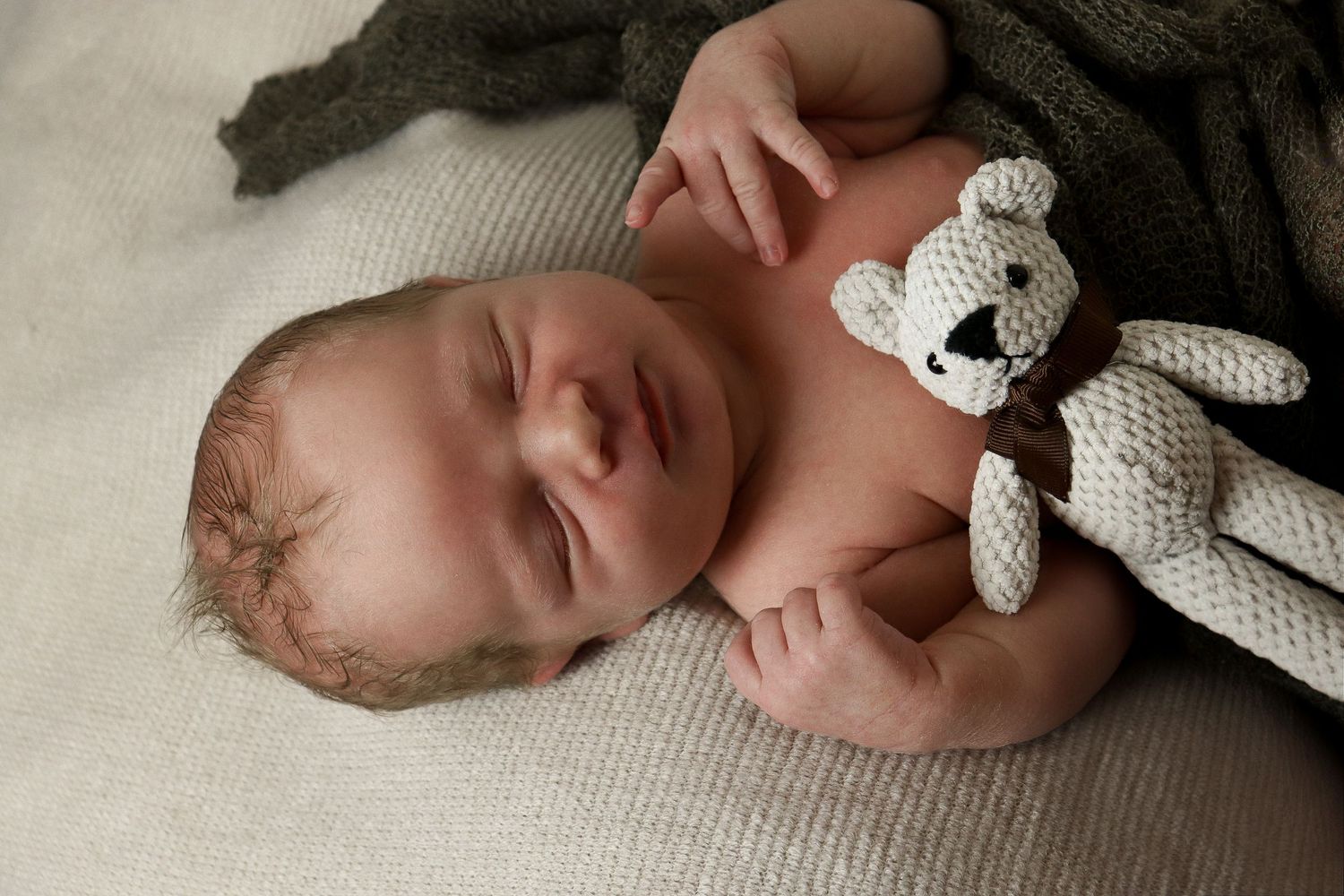 A newborn baby sleeps peacefully in a knit blanket while cuddling with a white teddy bear.