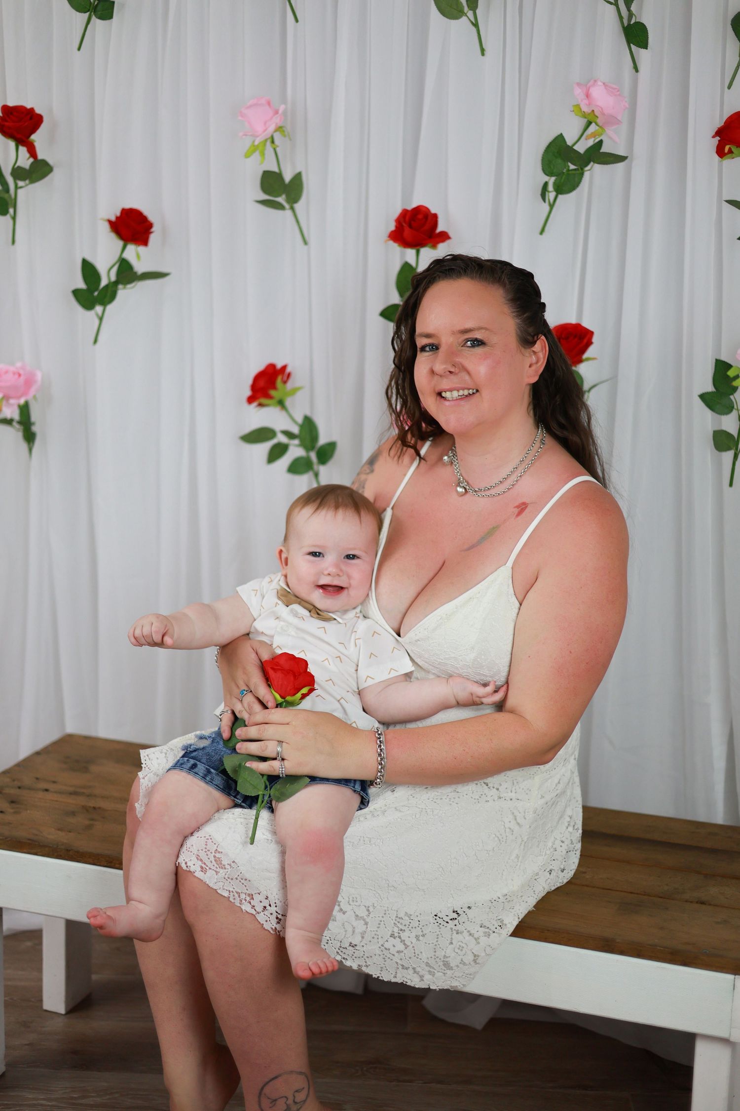 A parent and baby sit together on a wooden bench against a white curtain decorated with red and pink roses.