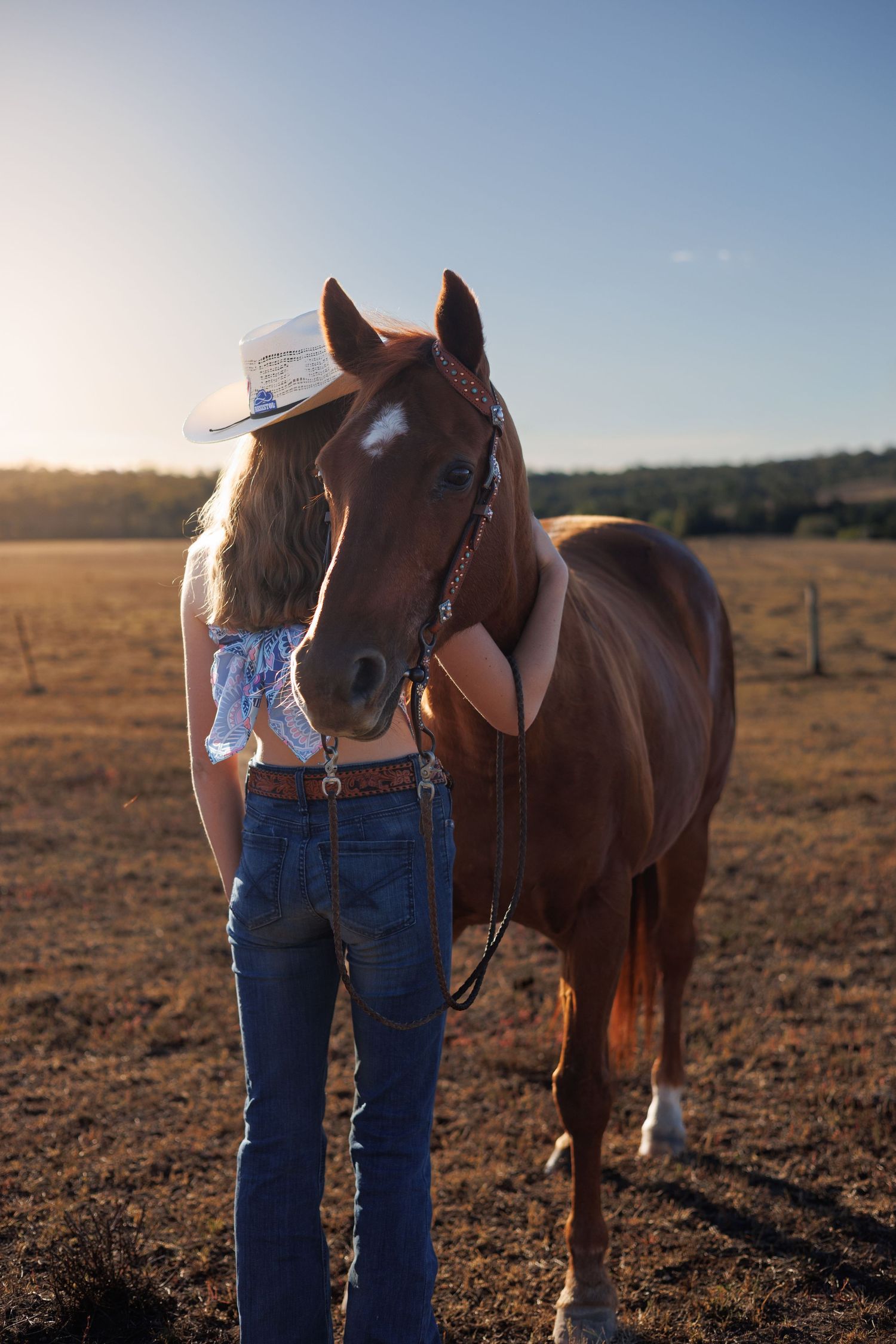 A young woman in jeans and cowboy hat stands with a chestnut horse in a golden sunlit field.