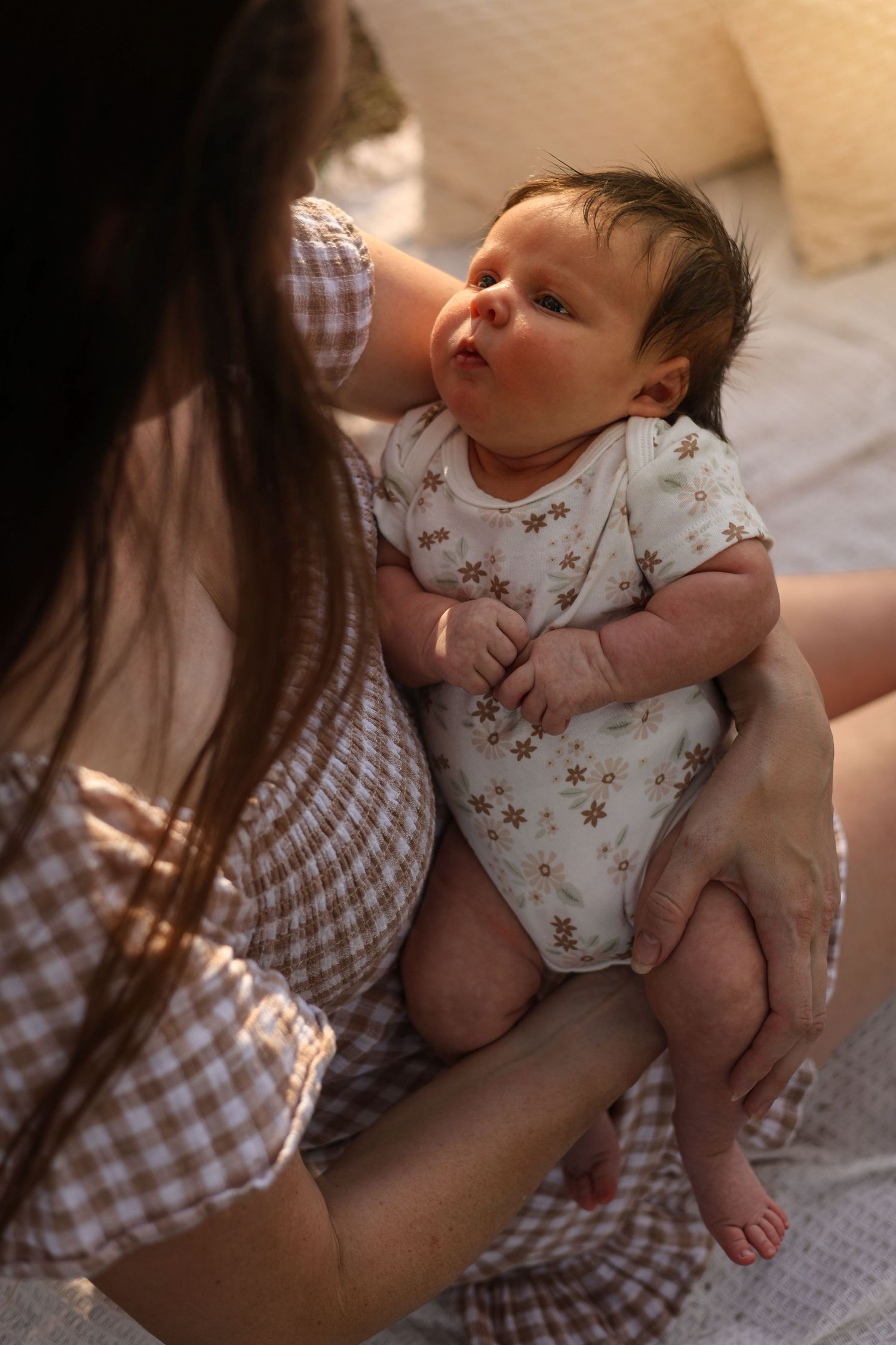 A mother in a gingham dress holds her newborn baby in a floral onesie in warm golden light.