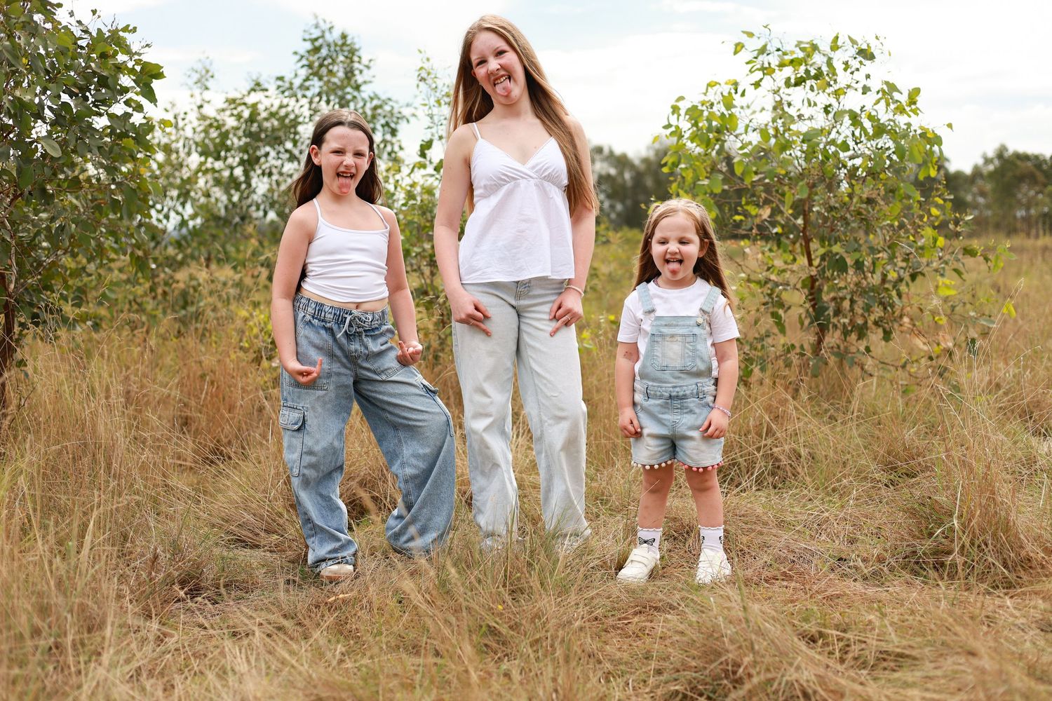Three girls in denim outfits smile outdoors in a grassy field surrounded by trees on a sunny day.