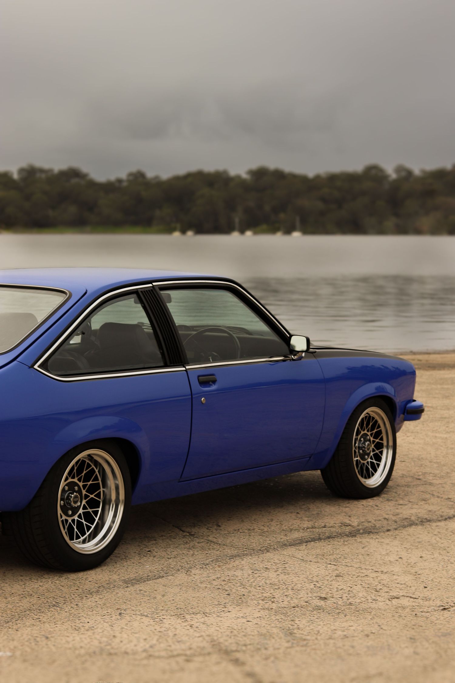 Classic blue torana with aftermarket wheels parked by a serene lakeside under cloudy skies.