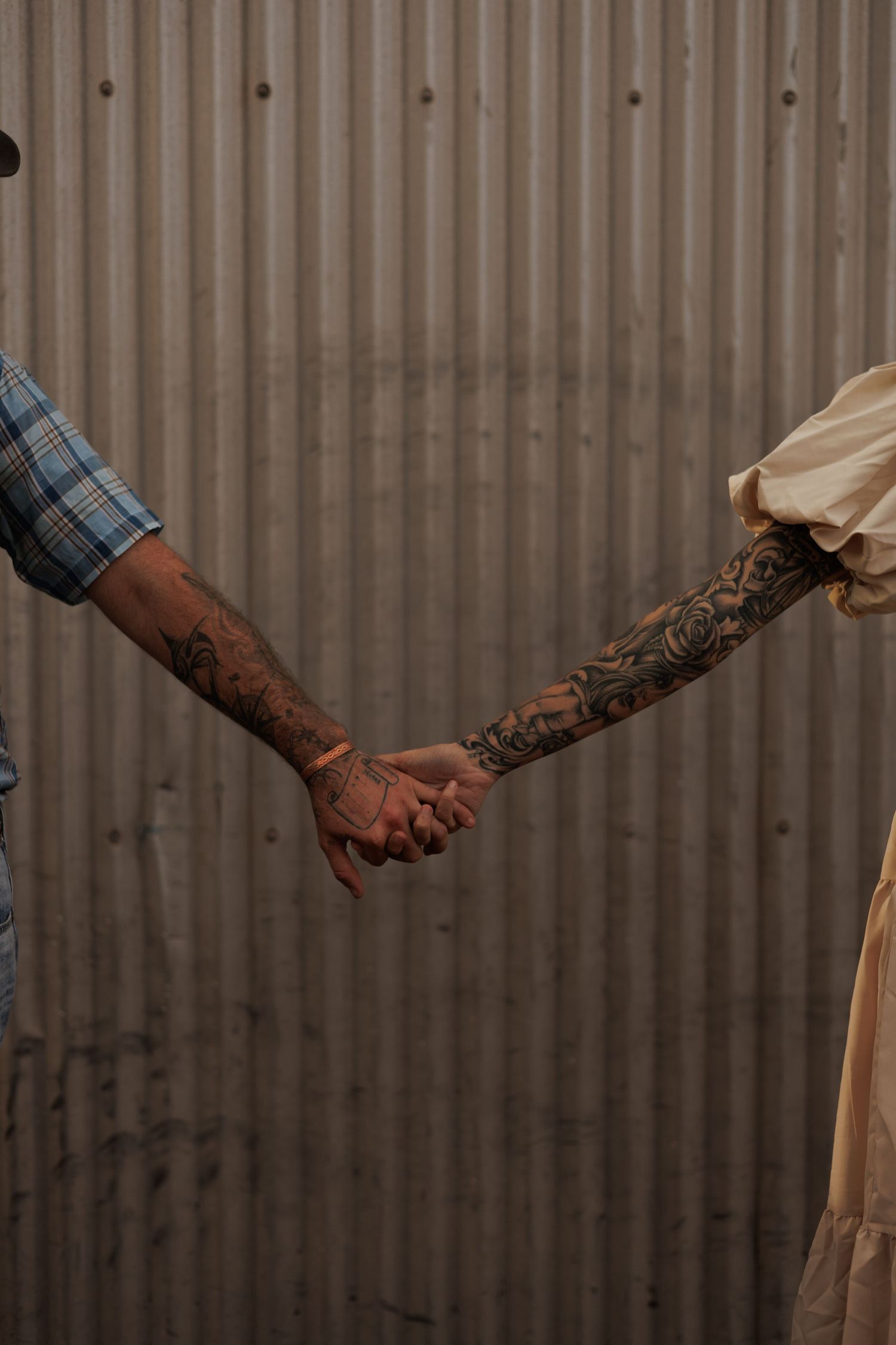 Two tattooed arms reaching toward each other, holding hands against a corrugated metal wall background.